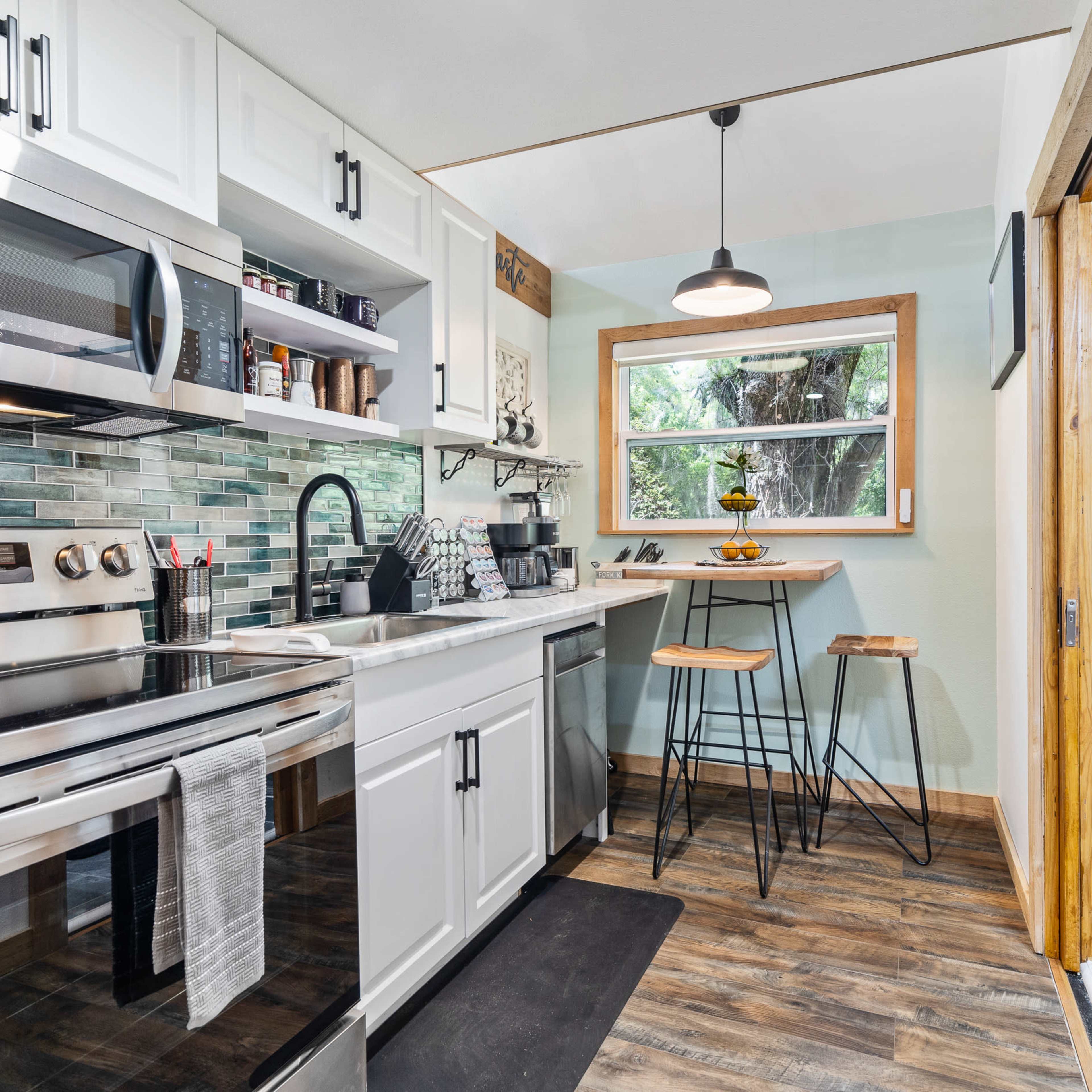 A compact kitchen featuring white cabinetry, stainless steel appliances, and a small dining area with two stools.