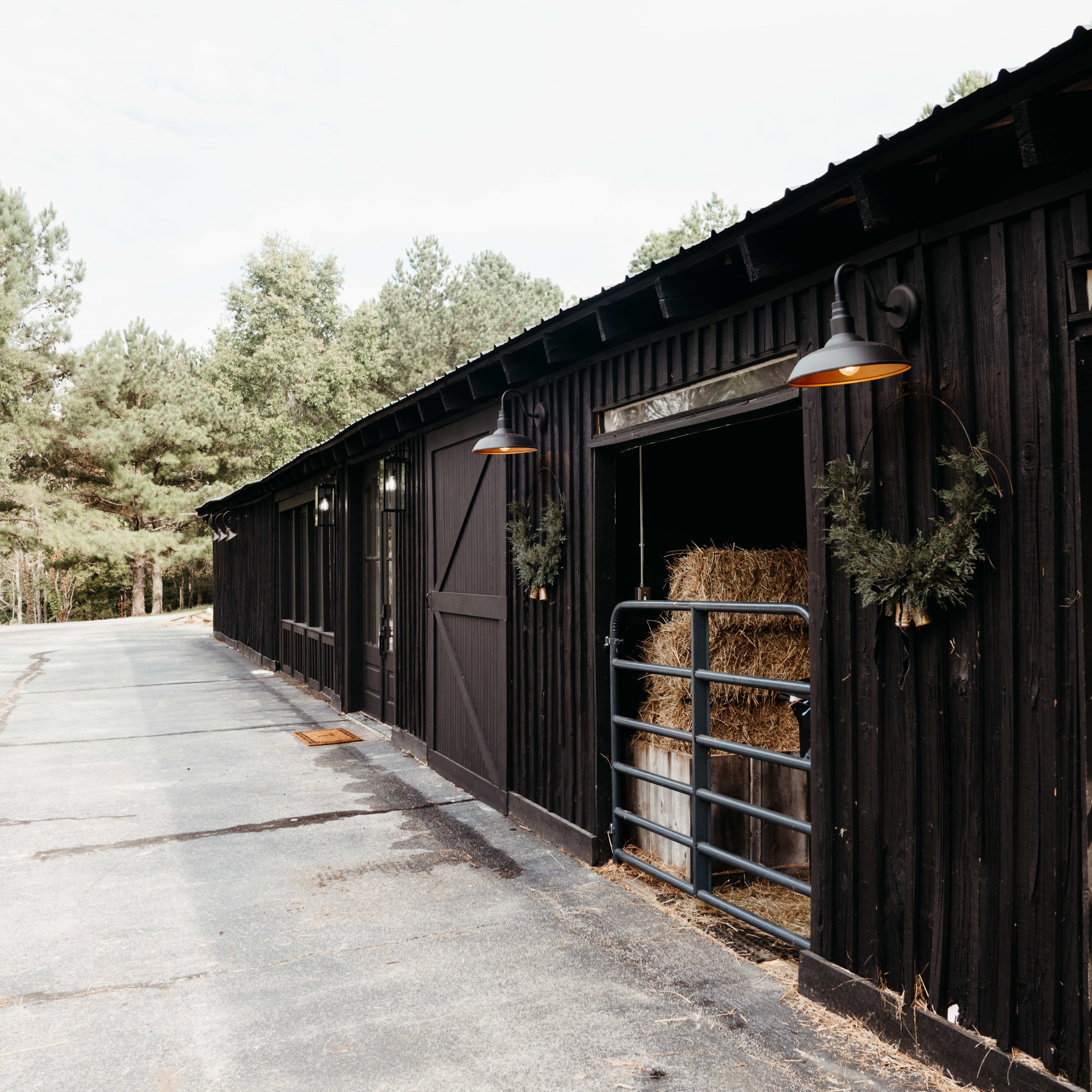 A black barn with open stalls displays bales of hay, lined along a paved path surrounded by trees.