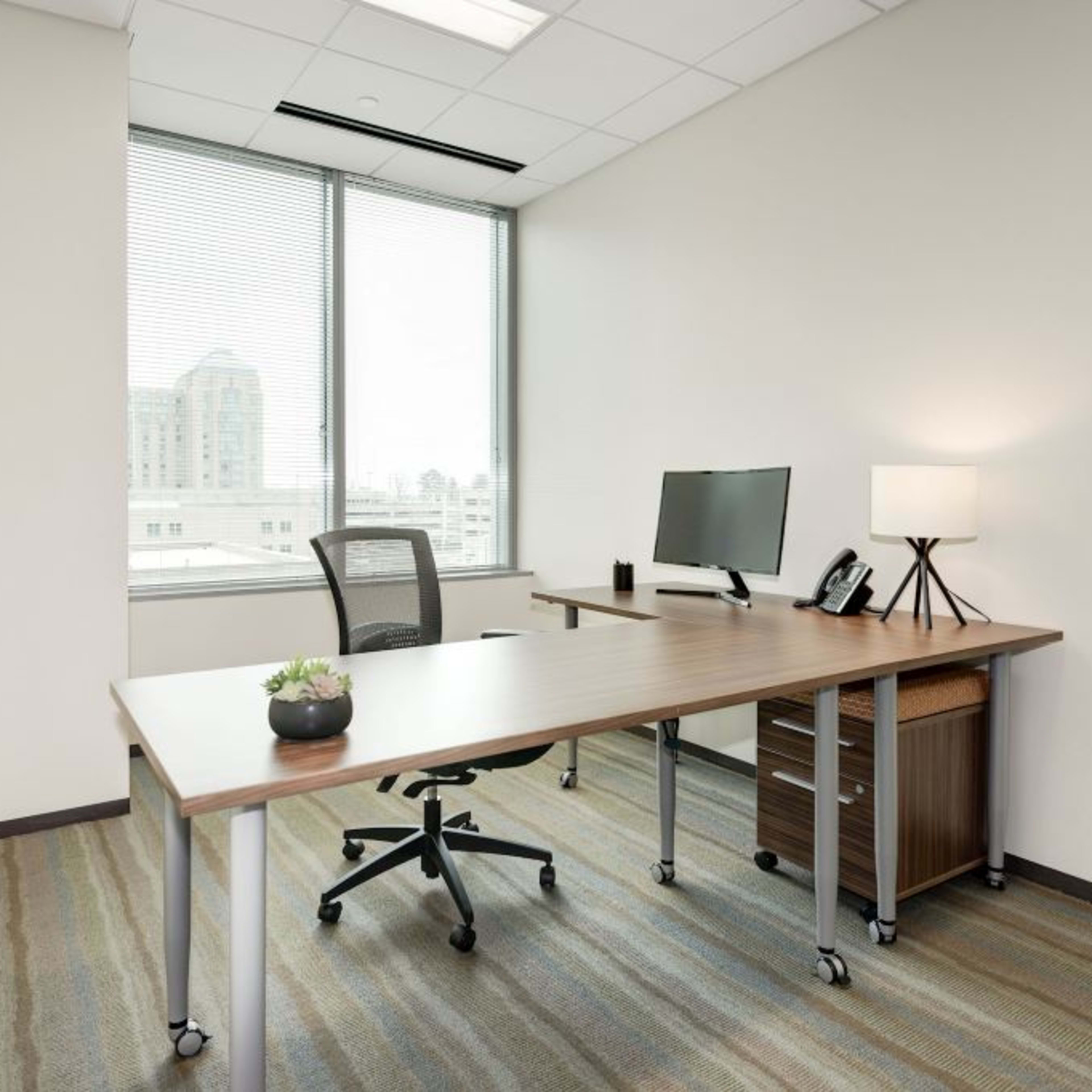 A modern office space featuring a wooden desk, an ergonomic chair, a computer monitor, a lamp, and a small plant on the desk, with large windows offering a view of a cityscape.