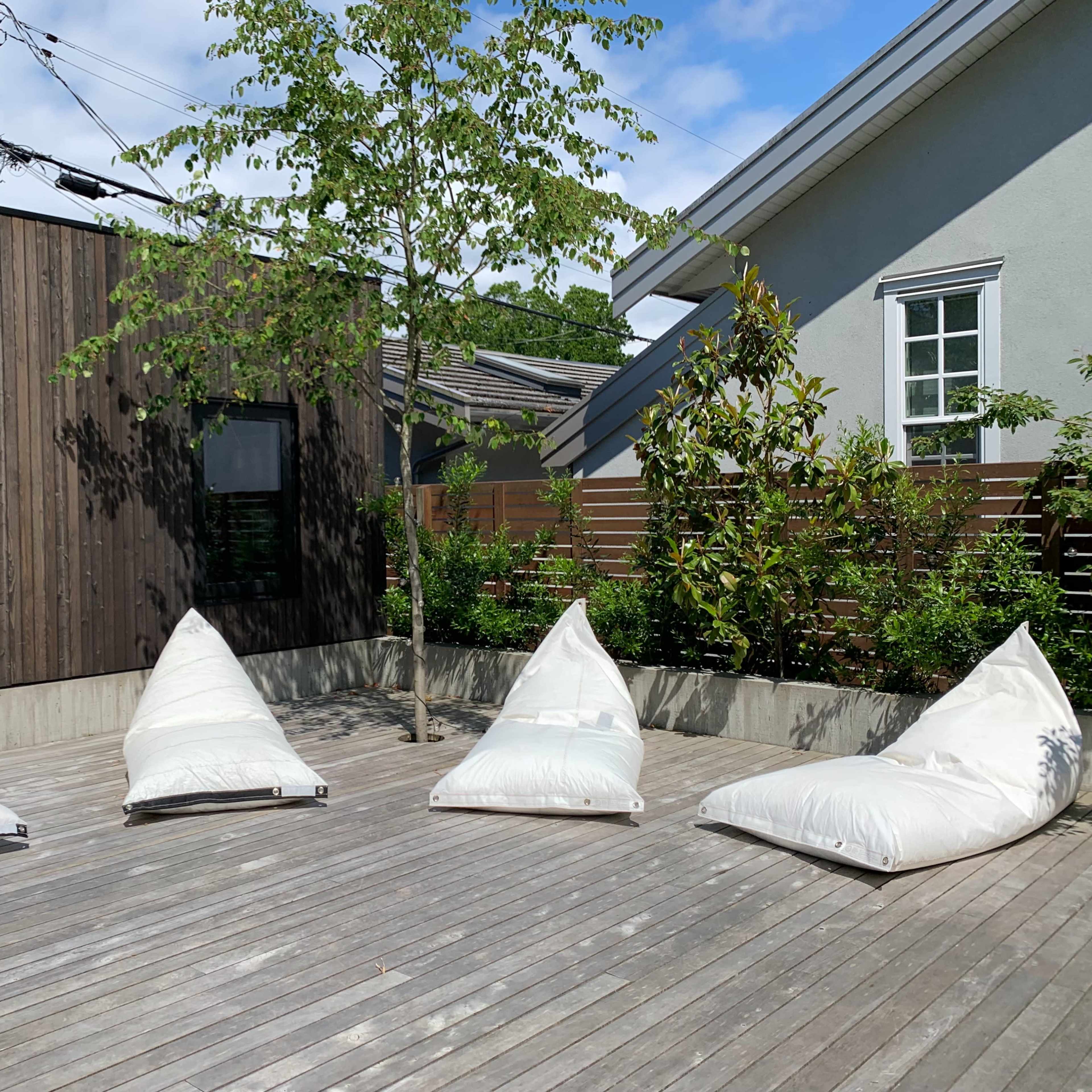 A wooden deck featuring four large, white bean bag chairs arranged around a small tree with greenery in the background.
