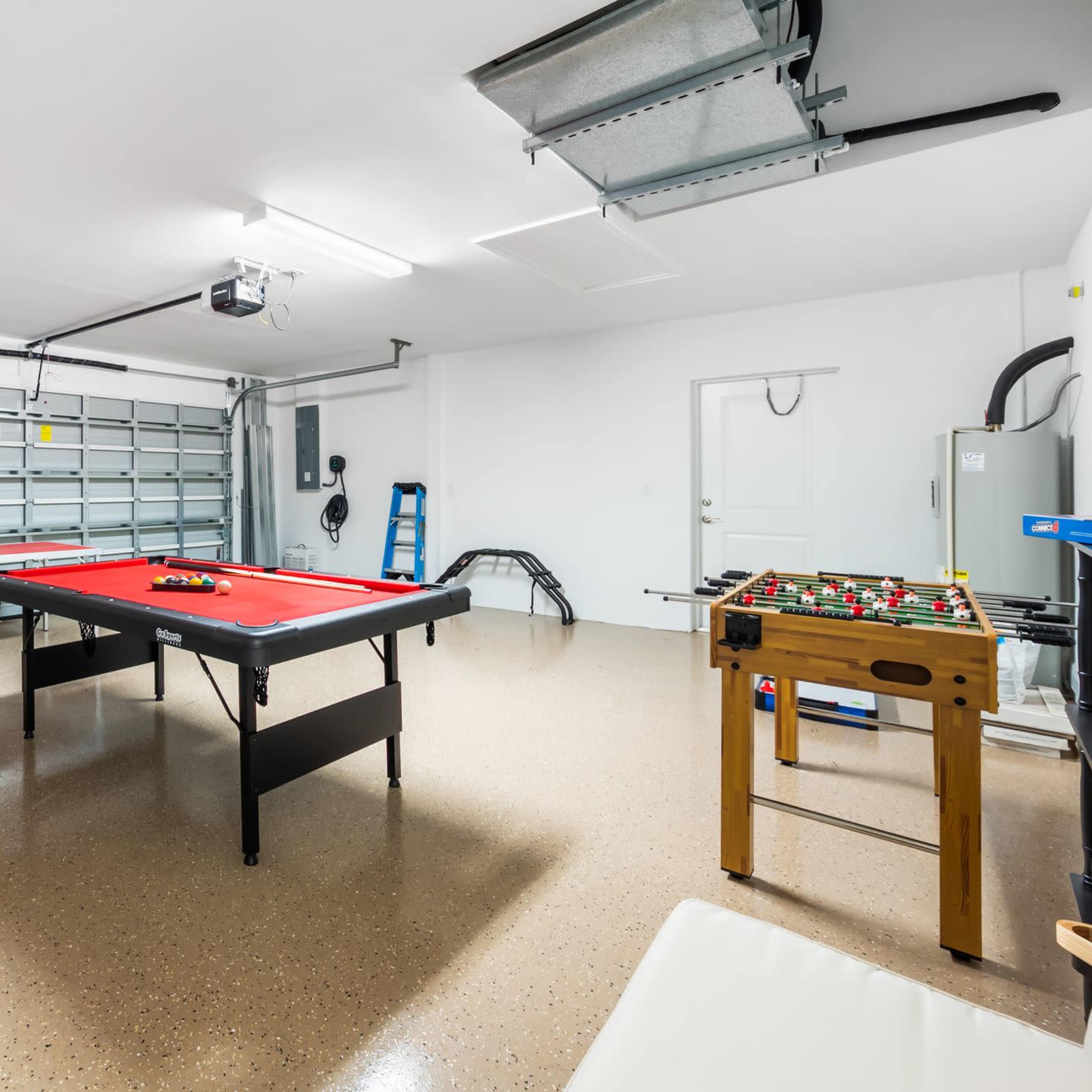 A spacious garage featuring a red pool table, a foosball table, and seating areas, with a neutral floor and overhead lighting.