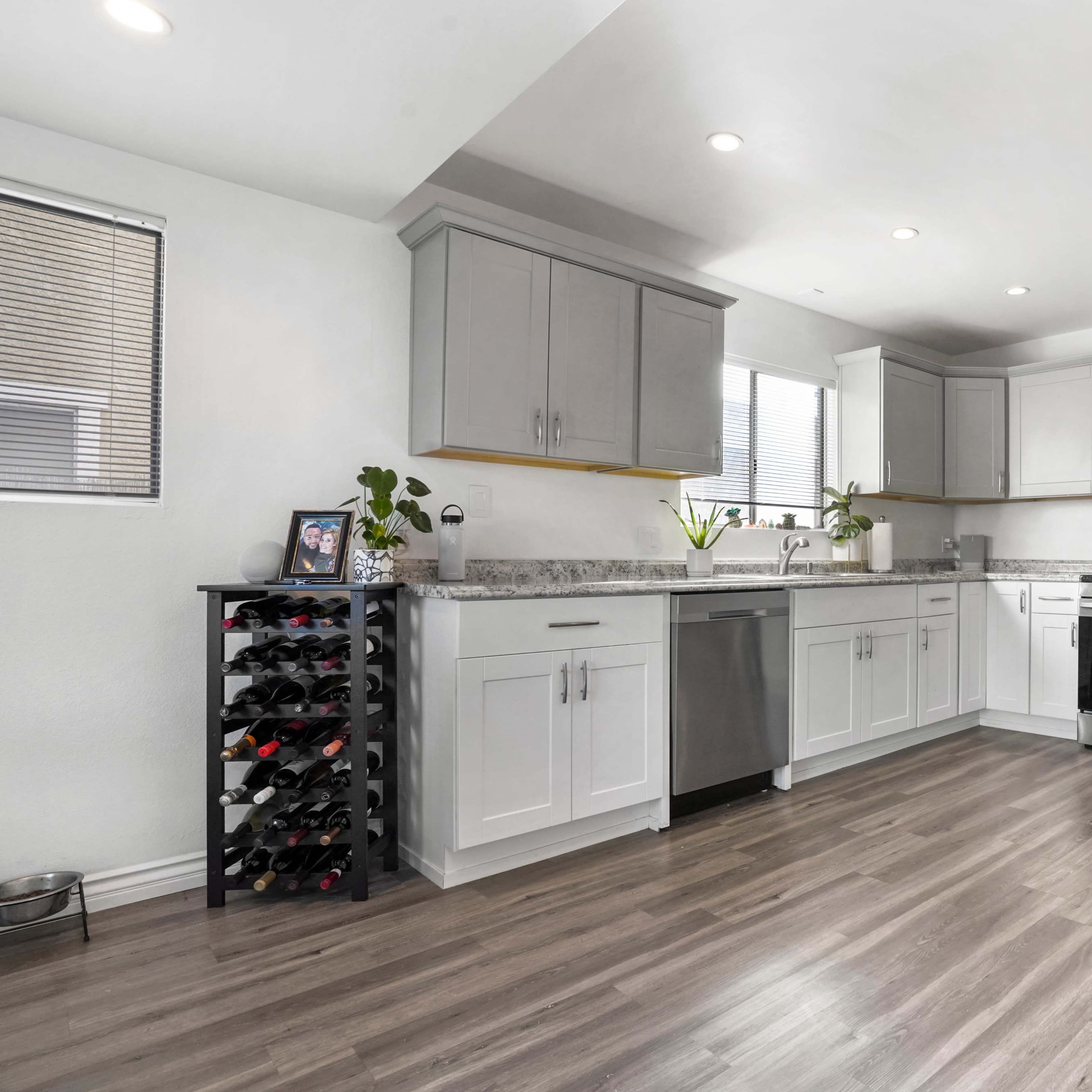 The modern kitchen features gray and white cabinetry, stainless steel appliances, a wine rack, and a spacious countertop with natural light coming through the windows.