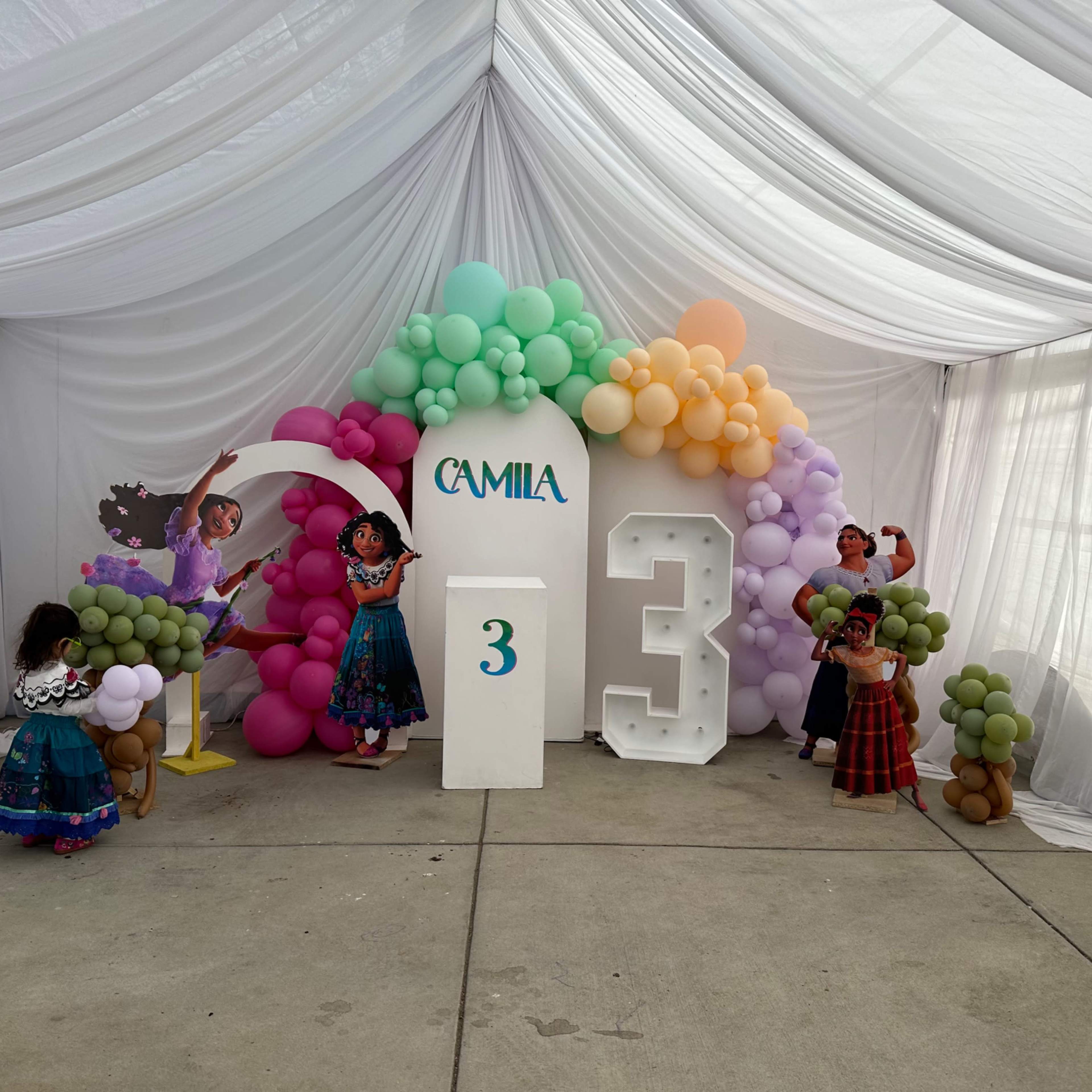 A festive birthday party setup with colorful balloons and decorations, featuring a cake display and the name "Camila" alongside the number "3."