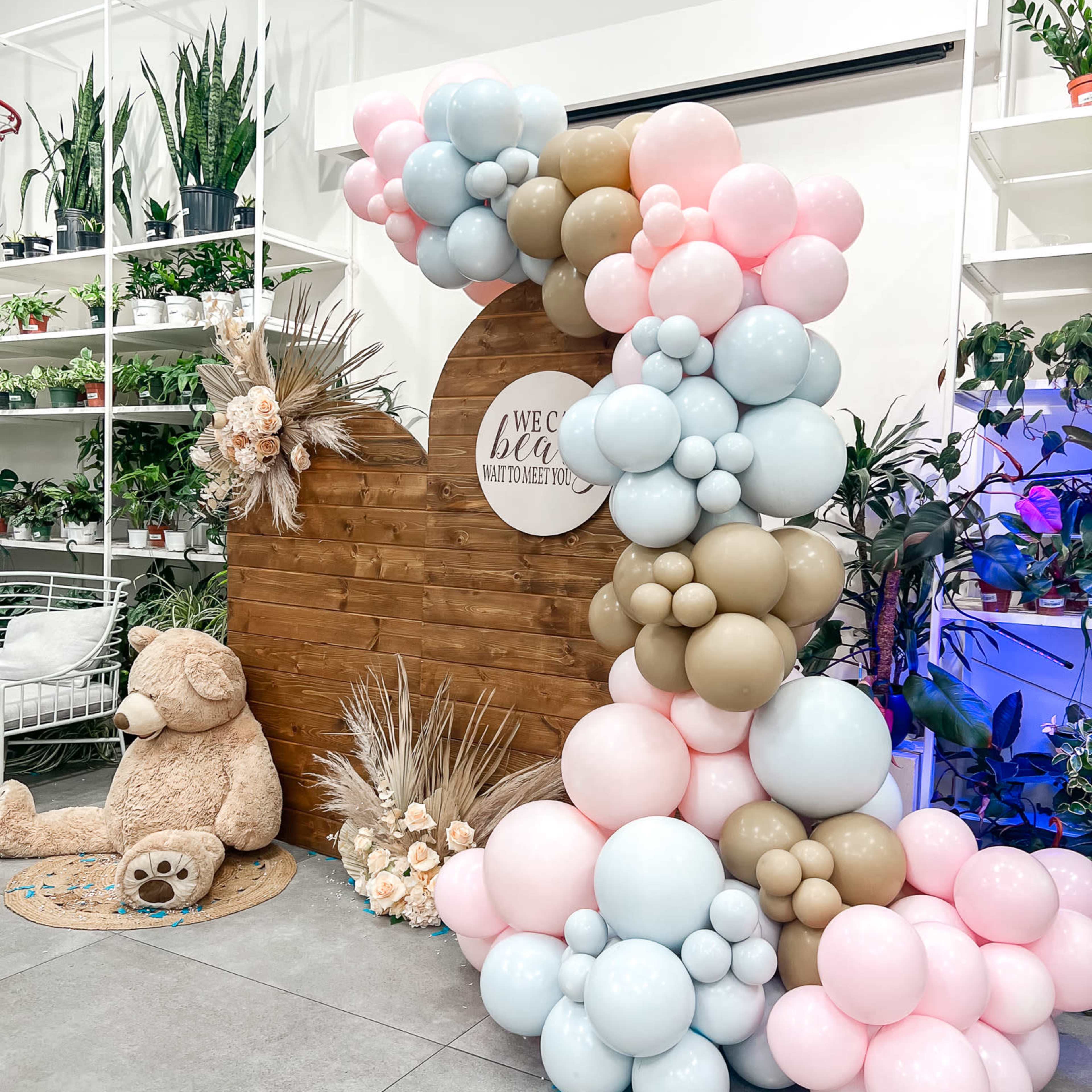 A decorative arch made of balloons in pastel colors frames a circular sign that reads "WE BIG WELCOME TO MEET YOU," with greenery and floral arrangements in the background.