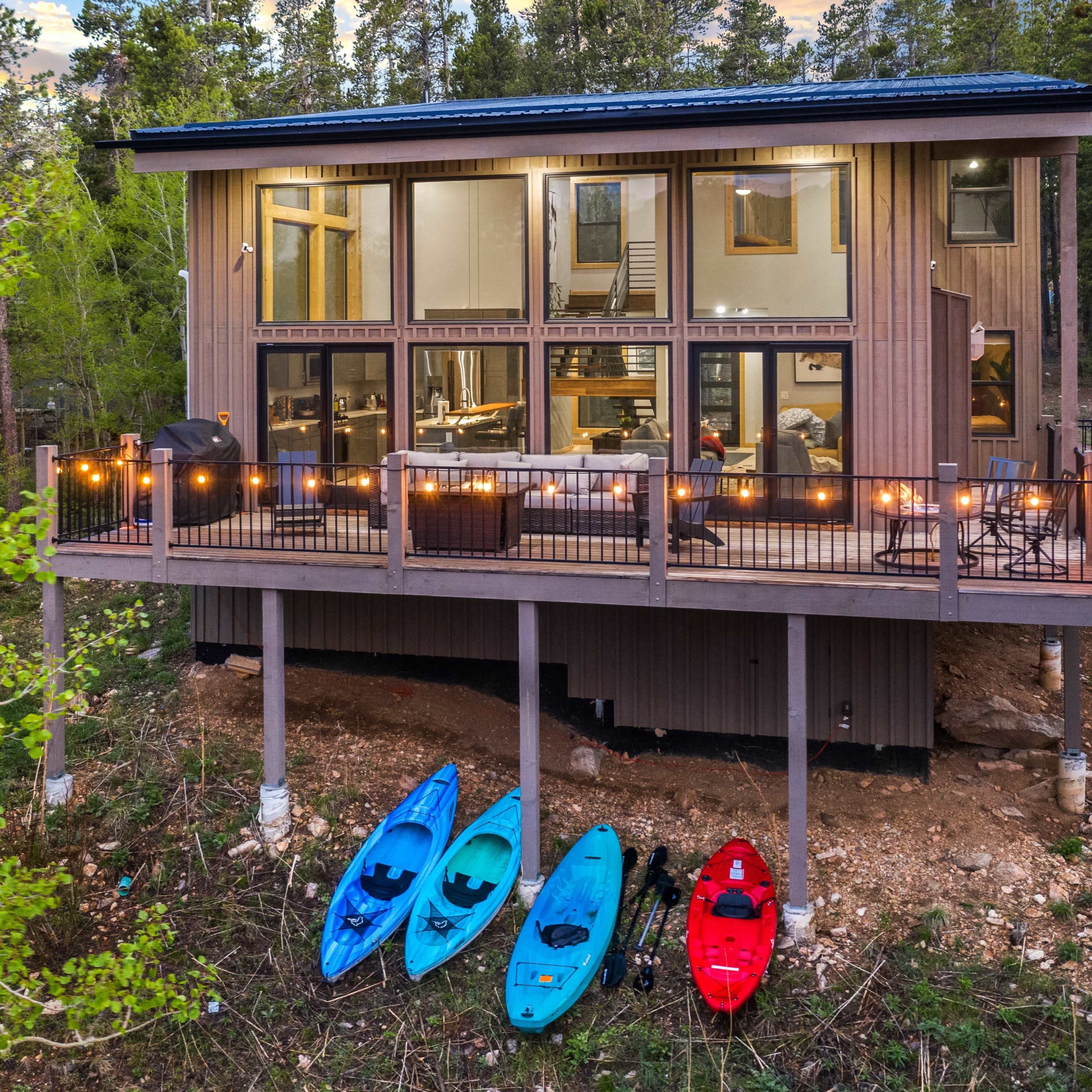 A modern cabin on stilts overlooks a wooded area, with three kayaks in blue and red parked on the ground below.