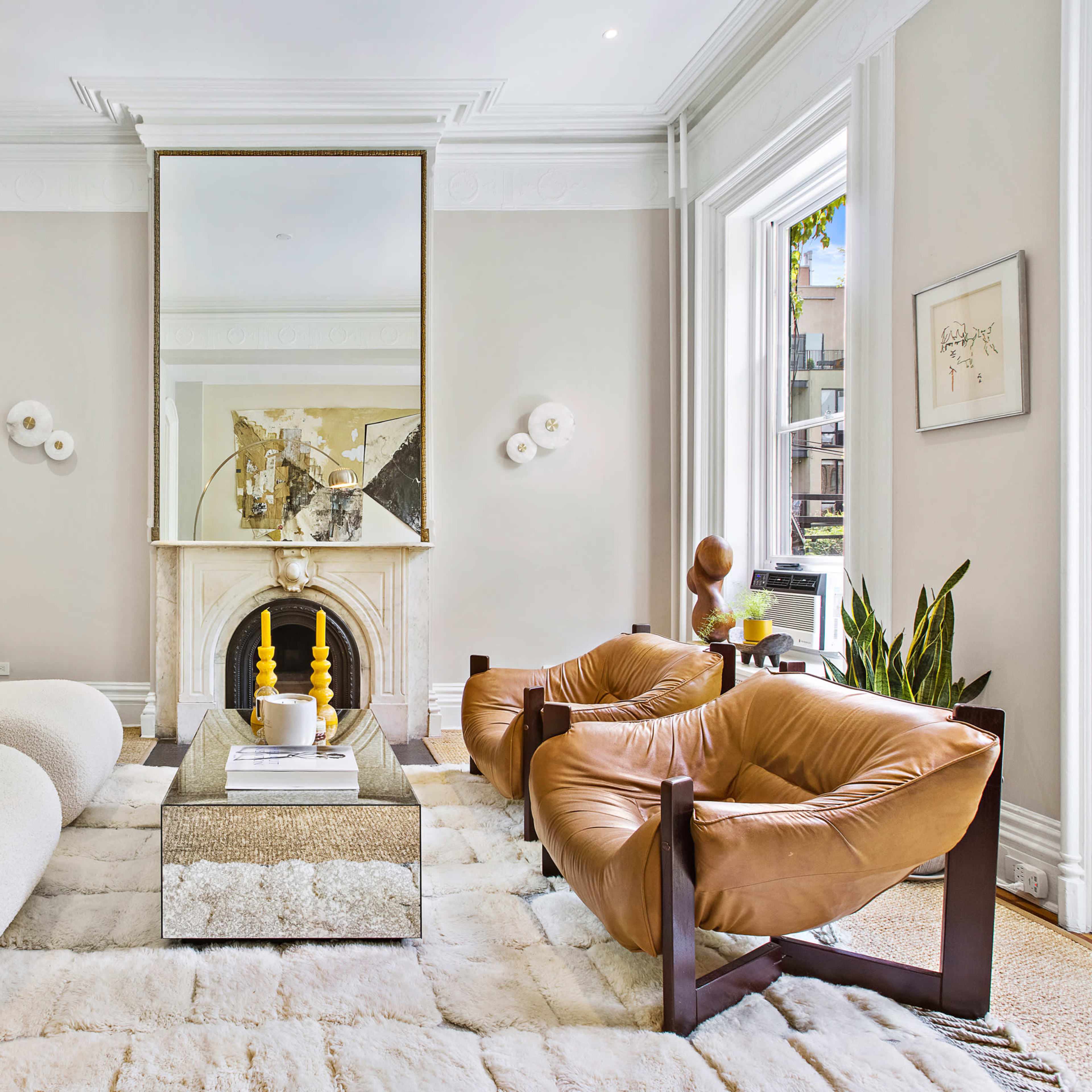 A modern living room with a plush white sofa, a brown leather chair, a large mirror, and a decorative coffee table in front of a fireplace.