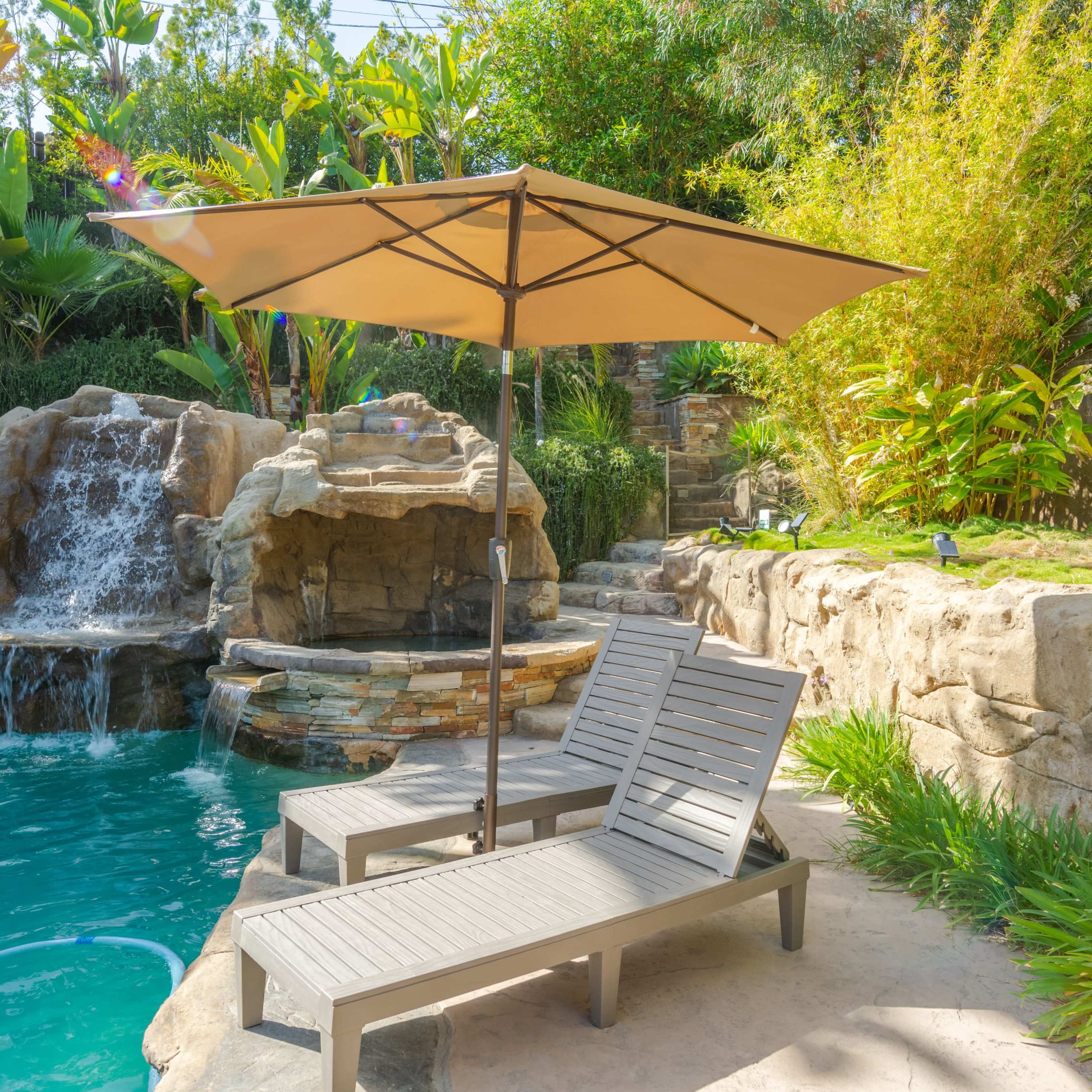 A tranquil poolside setting with two wooden lounge chairs positioned beneath a beige umbrella, adjacent to a waterfall and surrounded by lush greenery.