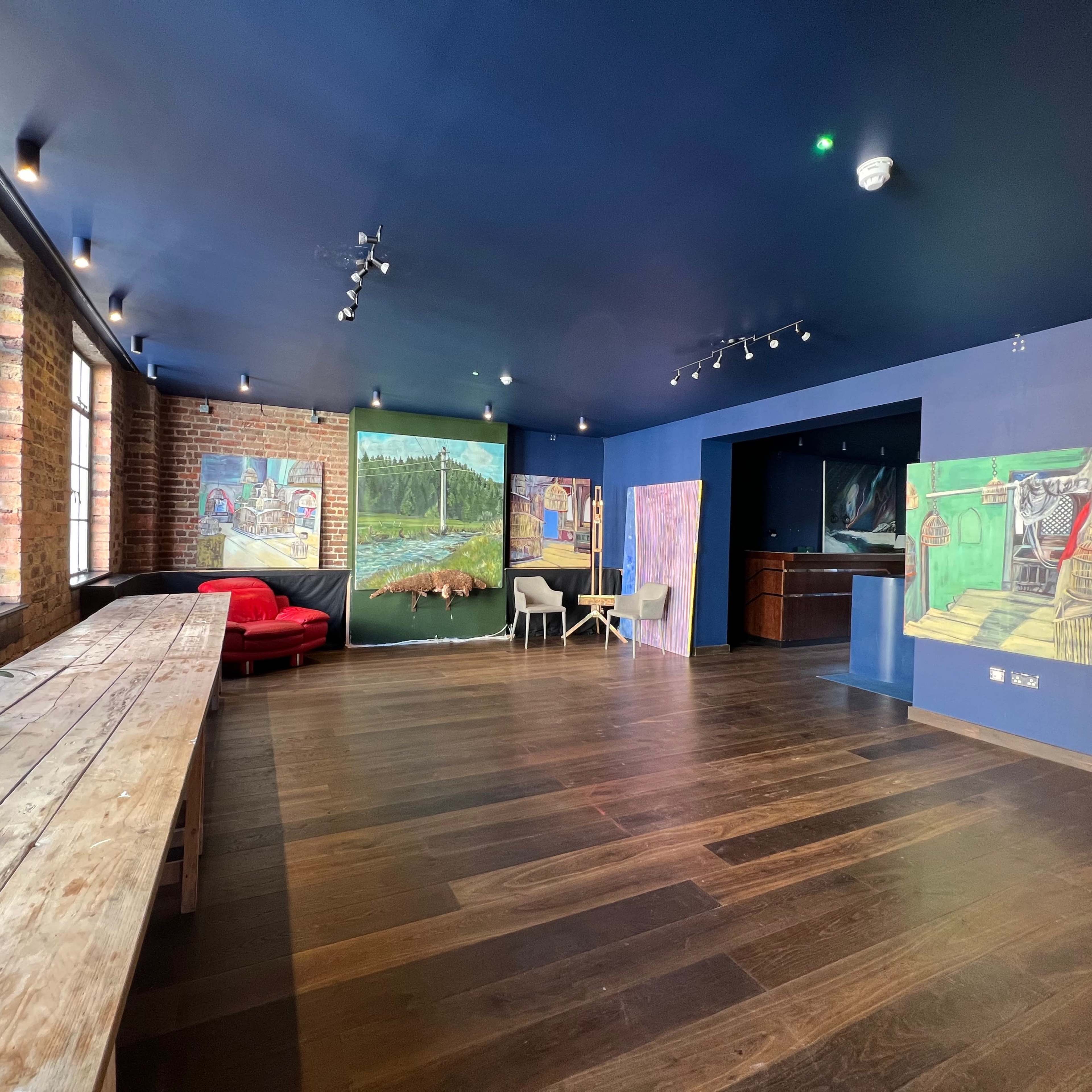 A spacious room with wooden flooring, large windows, and colorful murals on the walls, featuring a seating area and a long wooden table.