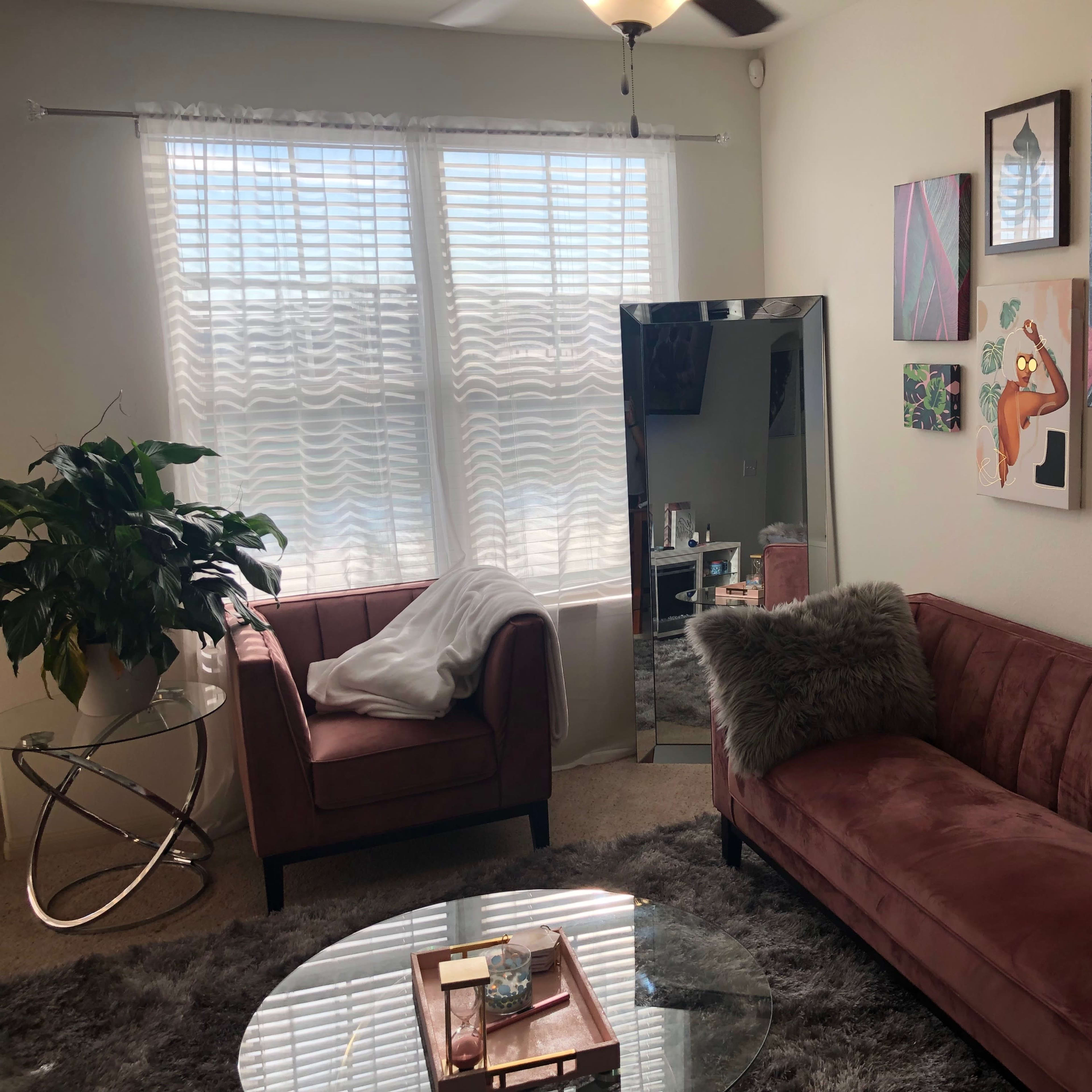 The scene features a living room with a pink velvet sofa, an armchair, a round glass coffee table, and various artworks displayed on the walls, illuminated by light from two windows.