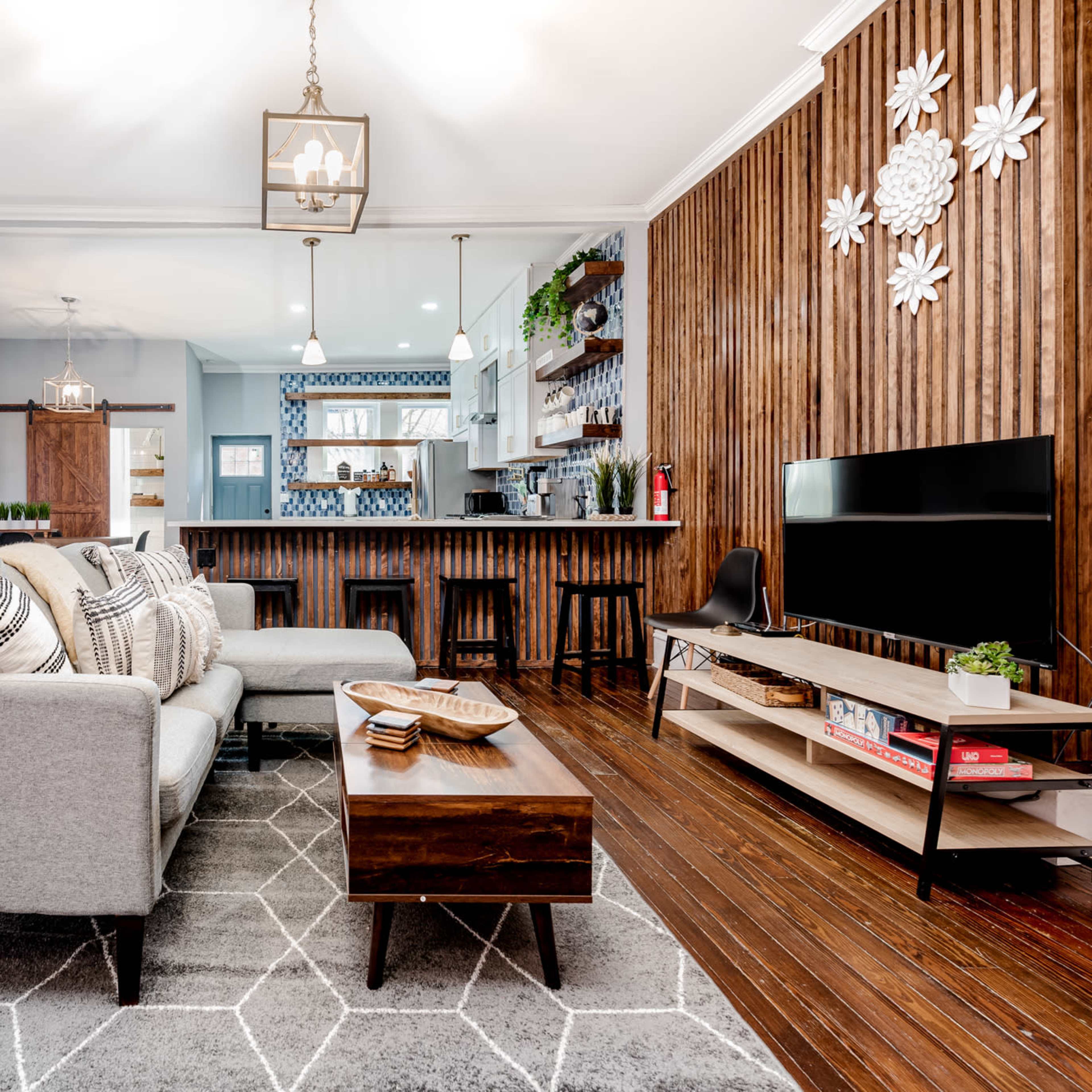 The living room features a gray sectional sofa, a wooden coffee table, and a television, with a backdrop of wood-paneled walls and a kitchen visible in the background.