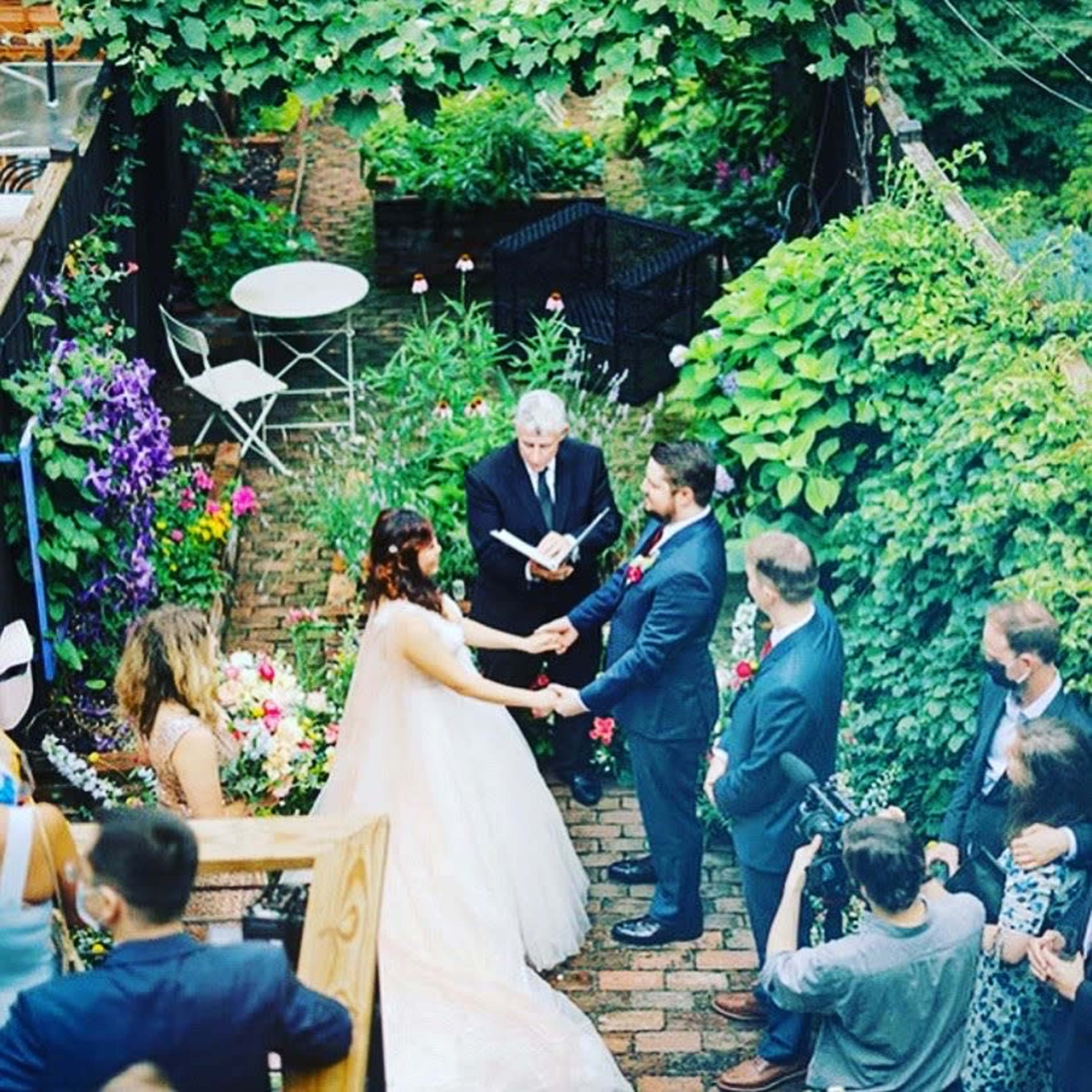 A couple stands hand-in-hand during their wedding ceremony in a garden setting, surrounded by flowers and guests.