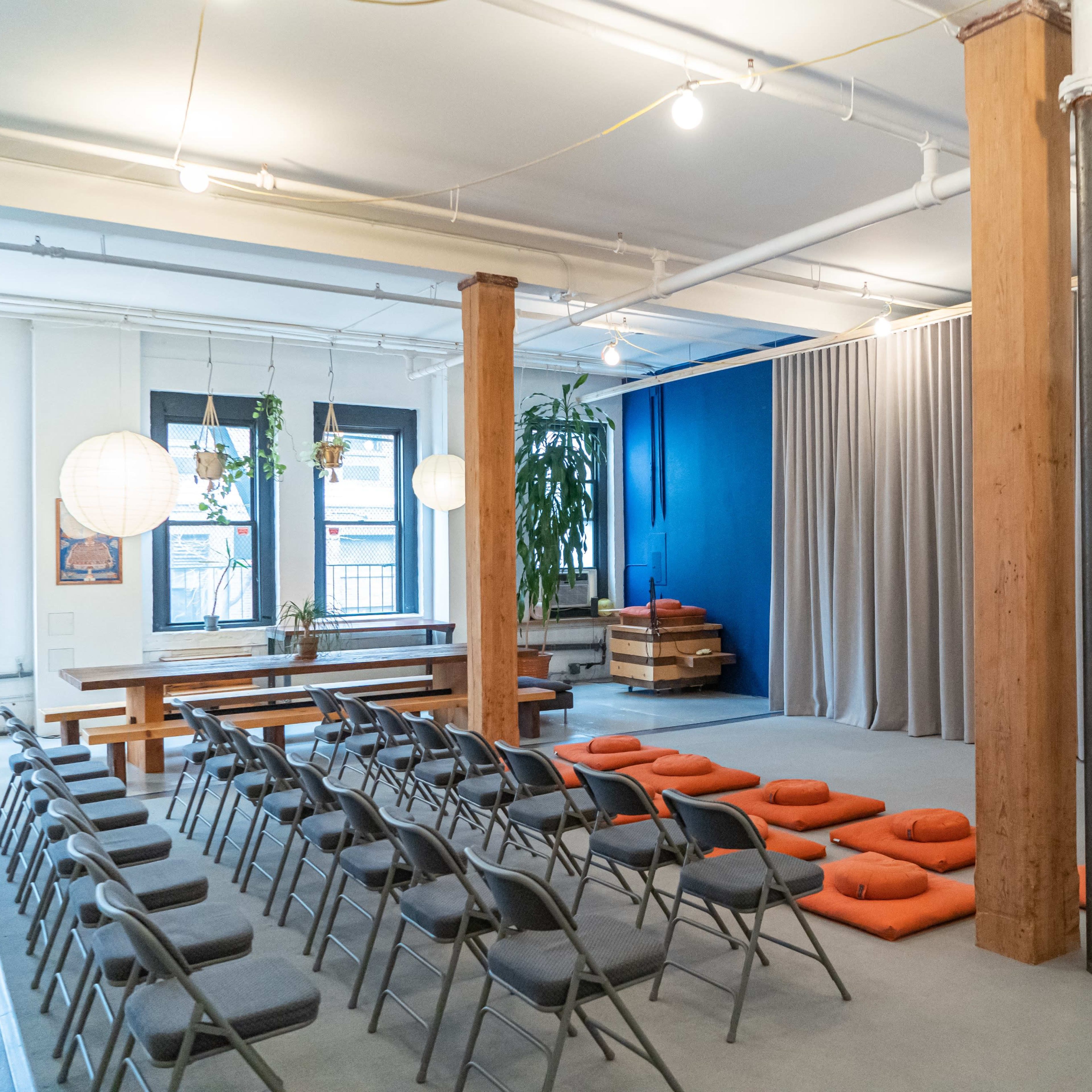 A spacious room with rows of folding chairs facing a curtain, orange cushions on the floor, and plants in the windows.
