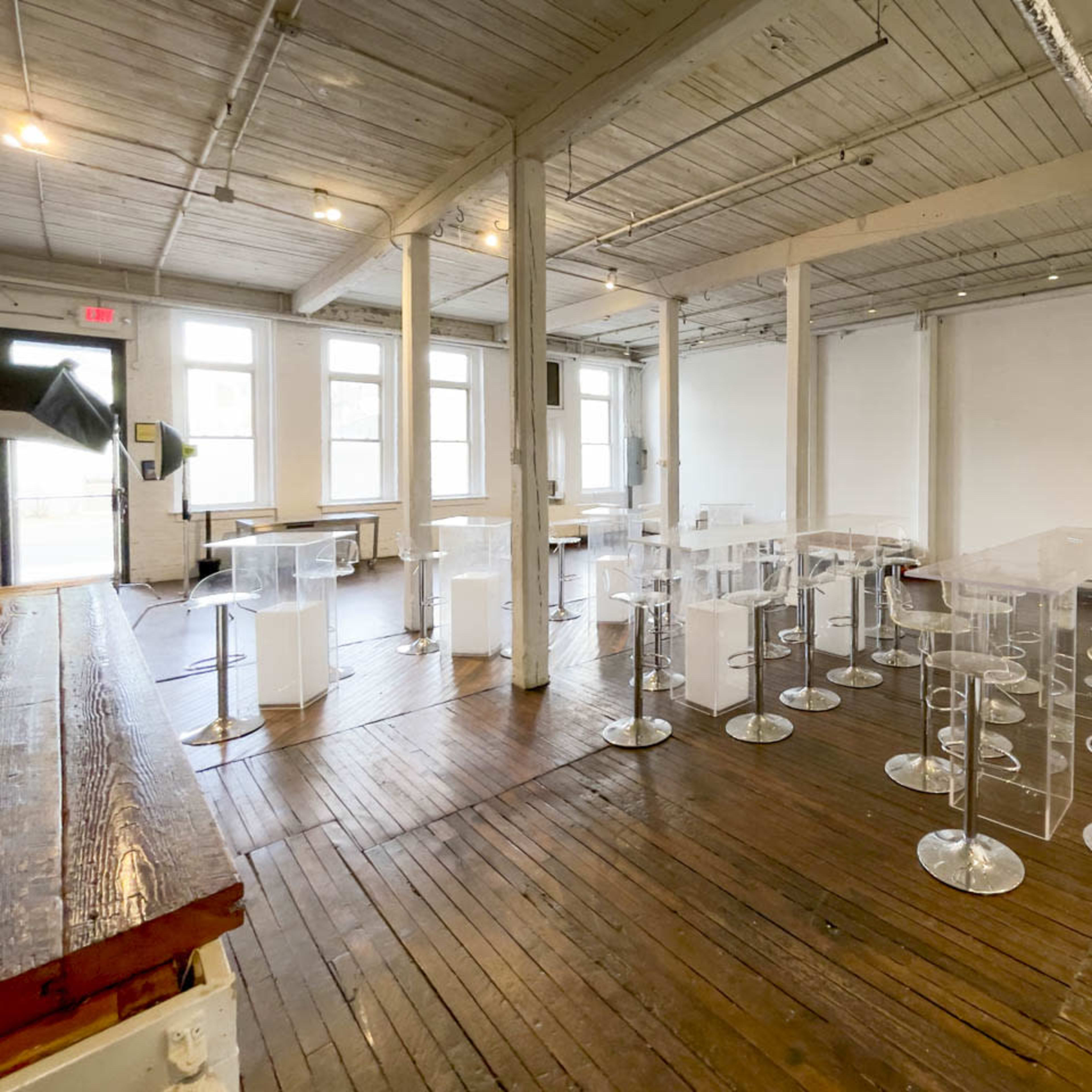 A spacious room with wooden floors, featuring transparent acrylic tables and stools arranged in a modern layout, alongside a few white seating areas.