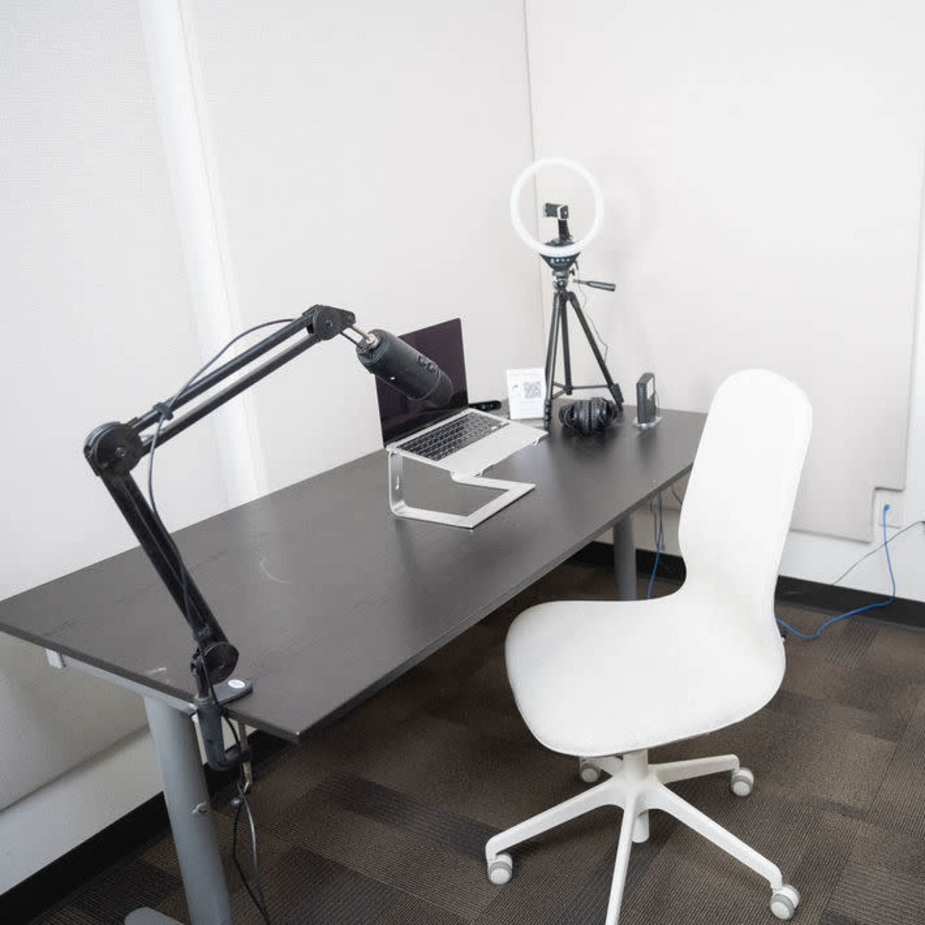 A minimalistic office space featuring a desk with a laptop, a microphone on an arm, a ring light, a white chair, and a potted plant.