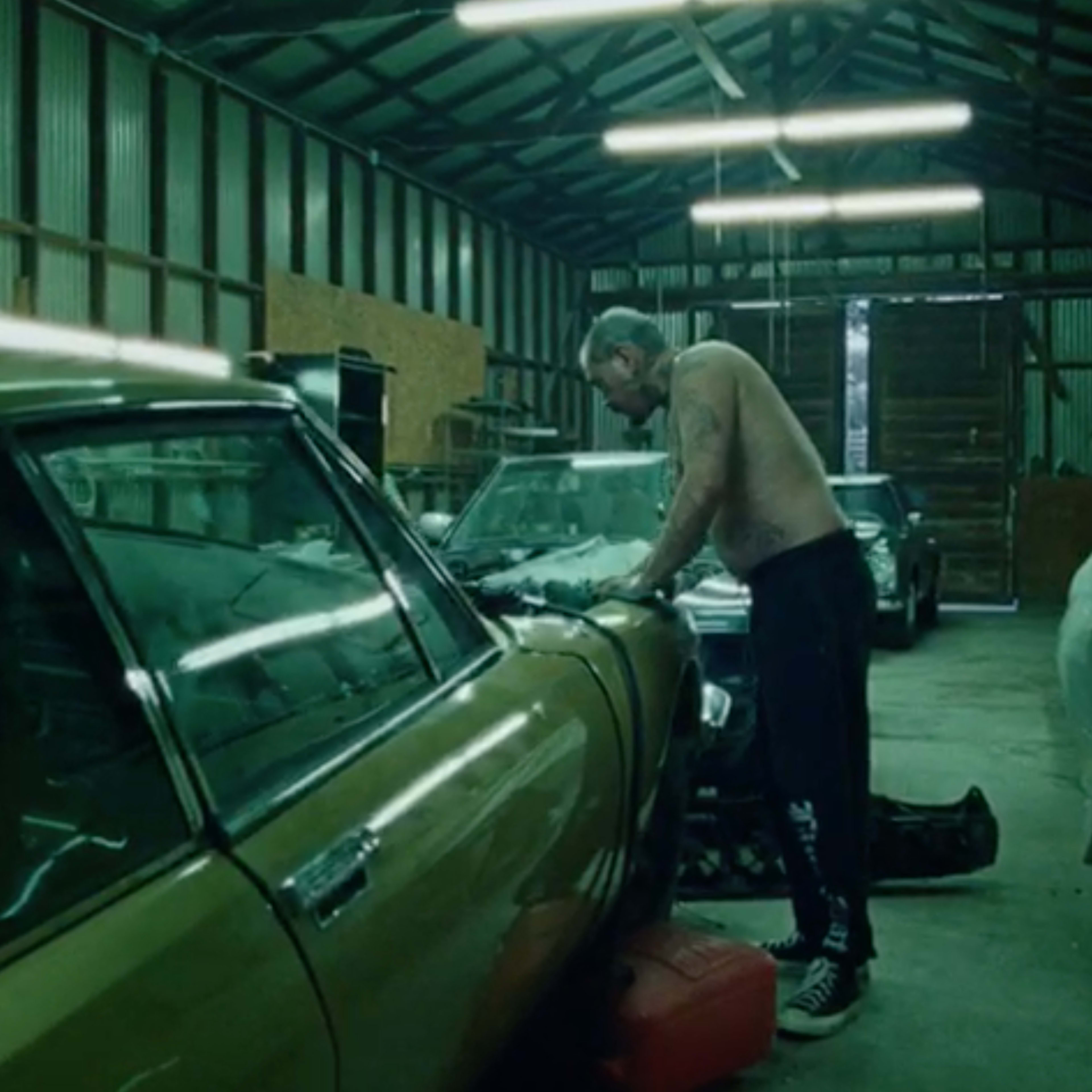 A shirtless man works on a classic car inside a dimly lit garage filled with various vehicles and tools.