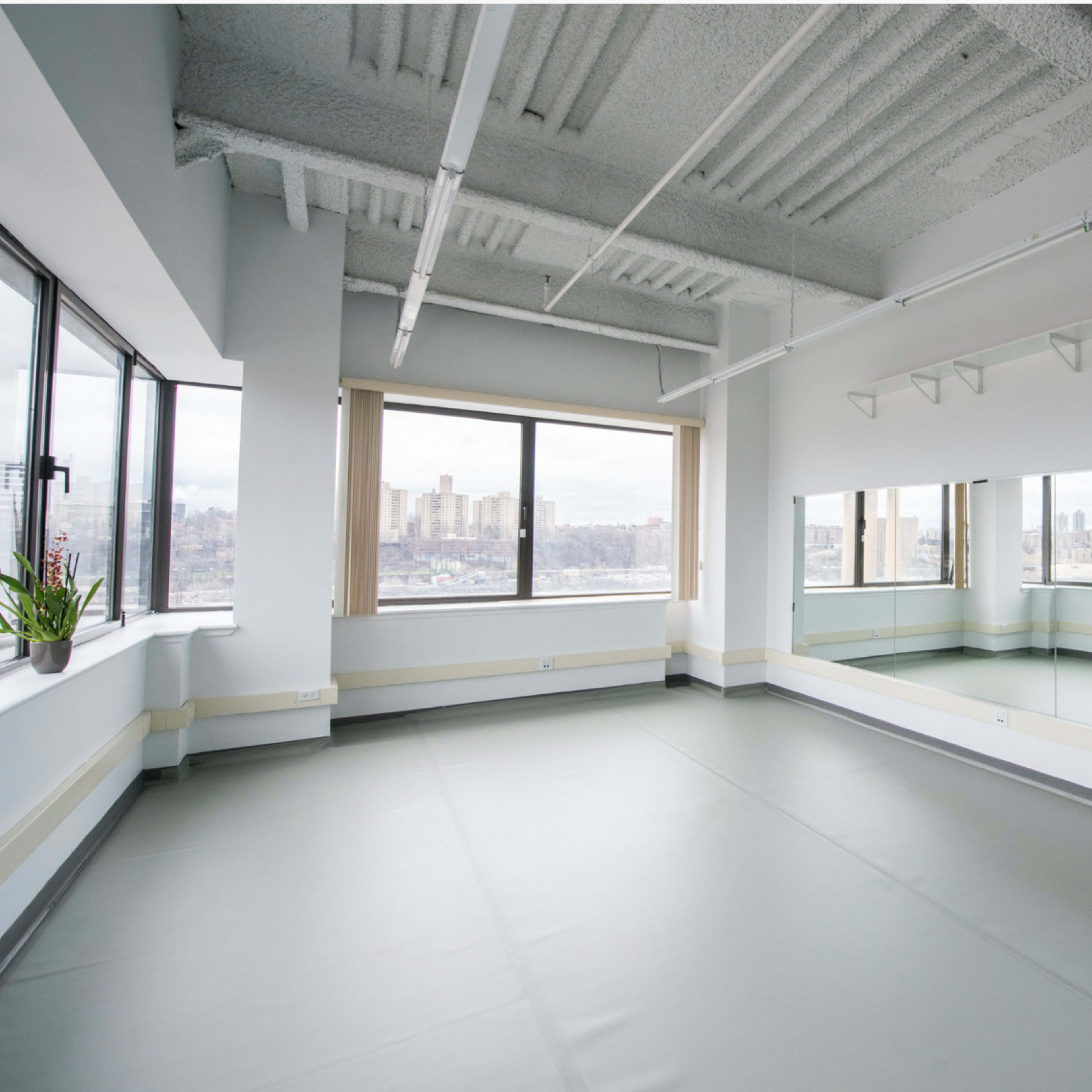 An empty dance studio with large windows, a mirrored wall, and a smooth floor.