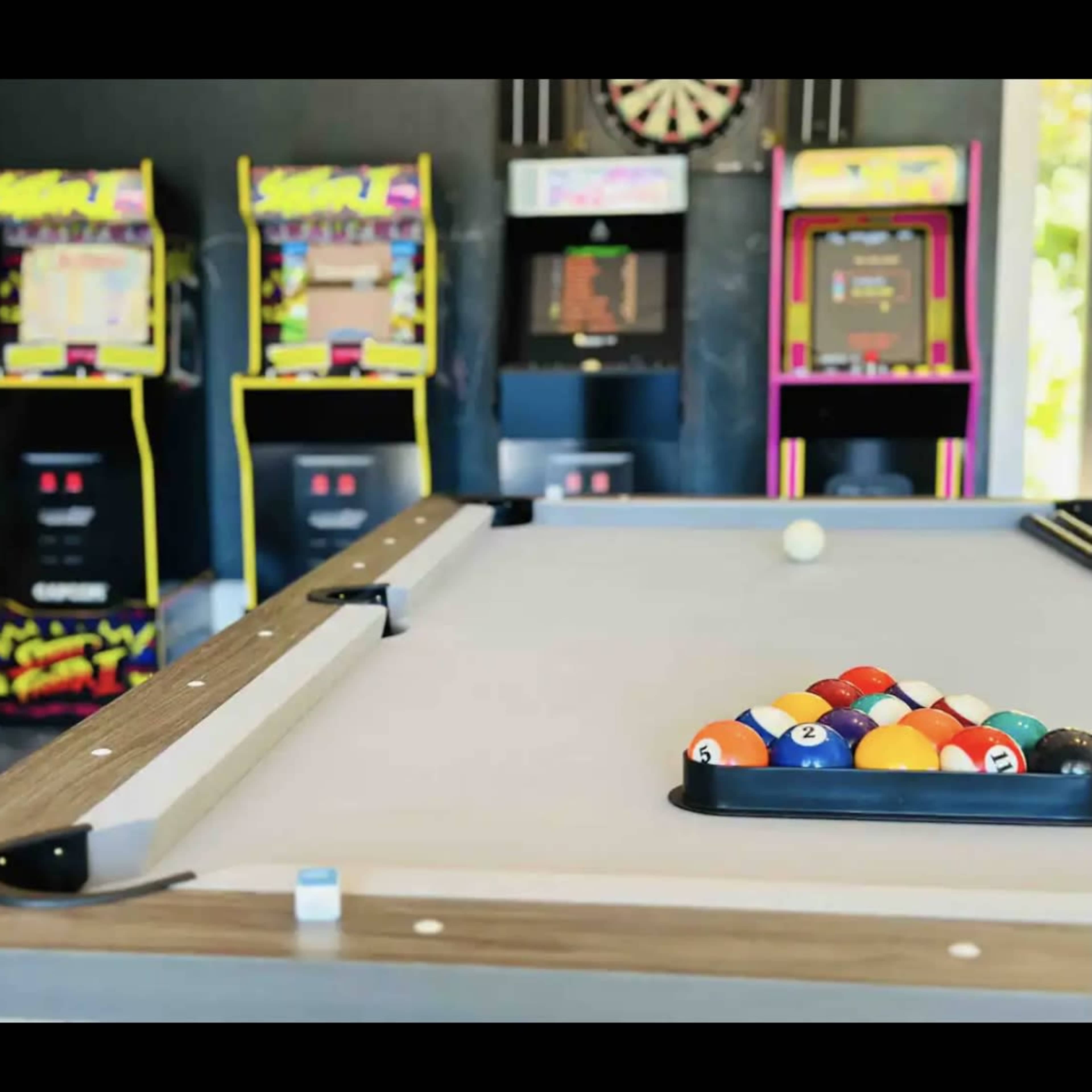 A billiard table in the foreground with a rack of colored balls and several vintage arcade machines in the background.