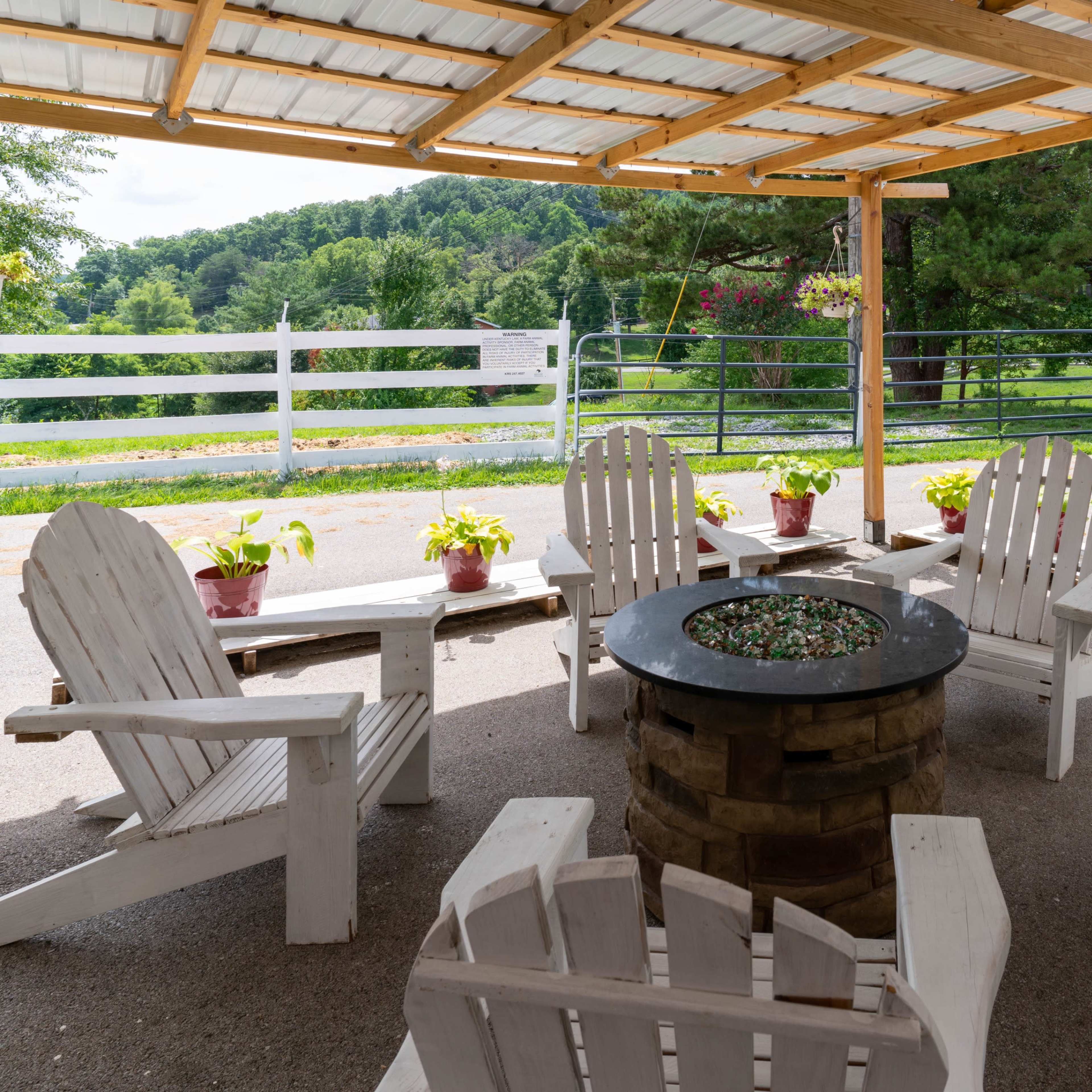 A sheltered seating area with white Adirondack chairs surrounds a stone fire pit, set against a backdrop of green hills and a white fence.