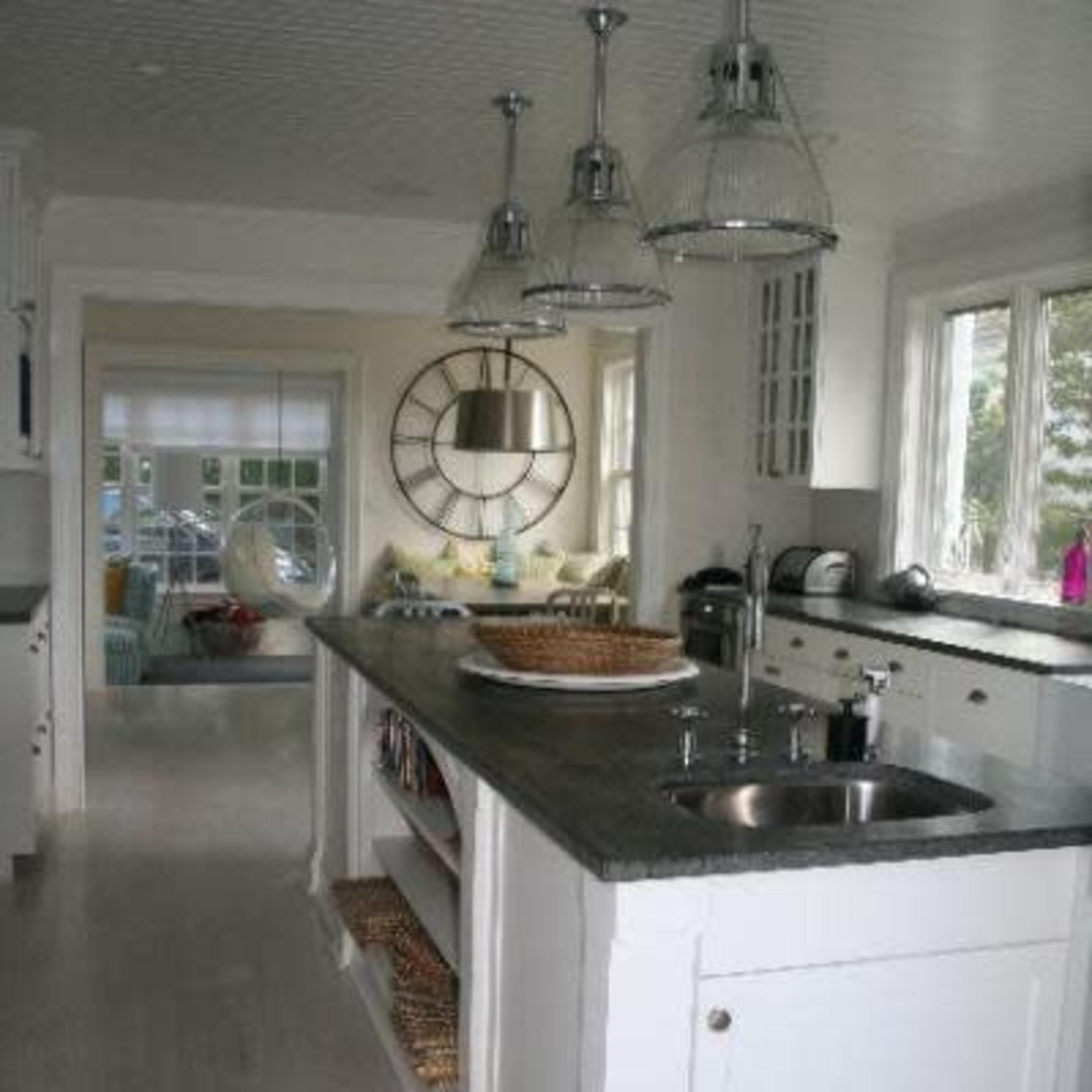 A modern kitchen with a central island, featuring pendant lights and a sink, leading to a brightly lit room with large windows.