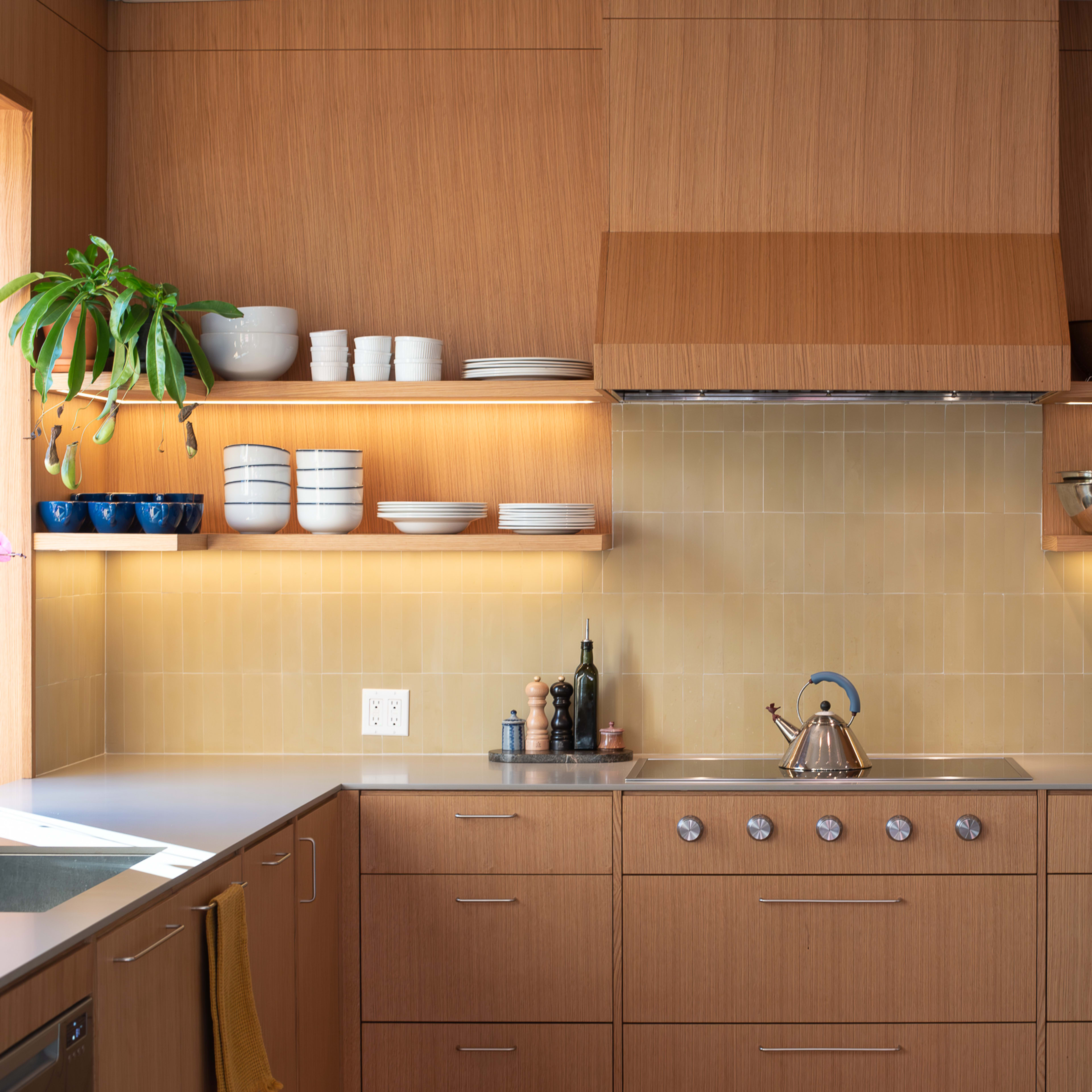 A modern kitchen with wooden cabinetry, open shelves displaying dishes, and a stove with a metallic kettle on the countertop.