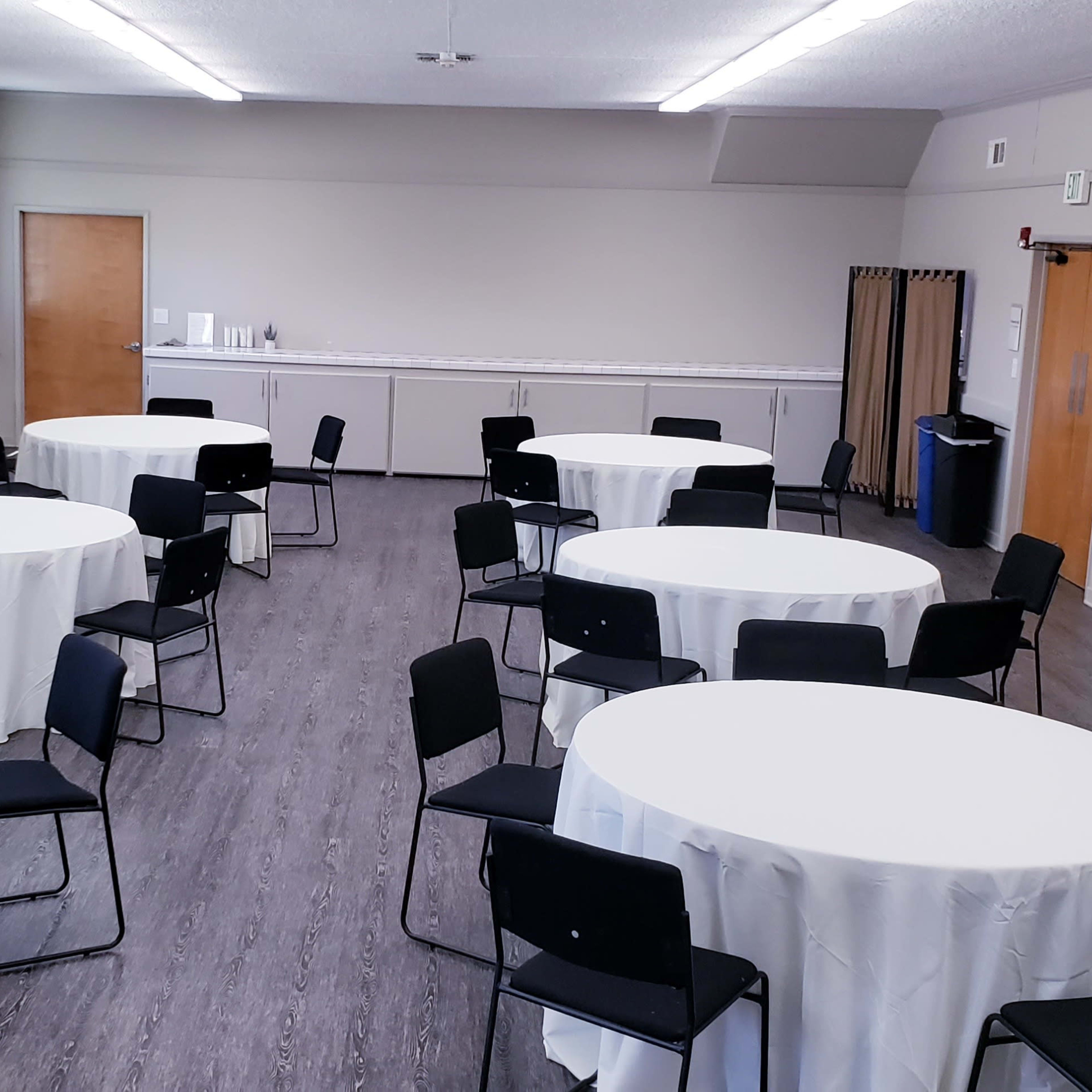 A room set up with multiple round tables covered with white tablecloths, surrounded by black chairs, and featuring plain walls and a few doors.