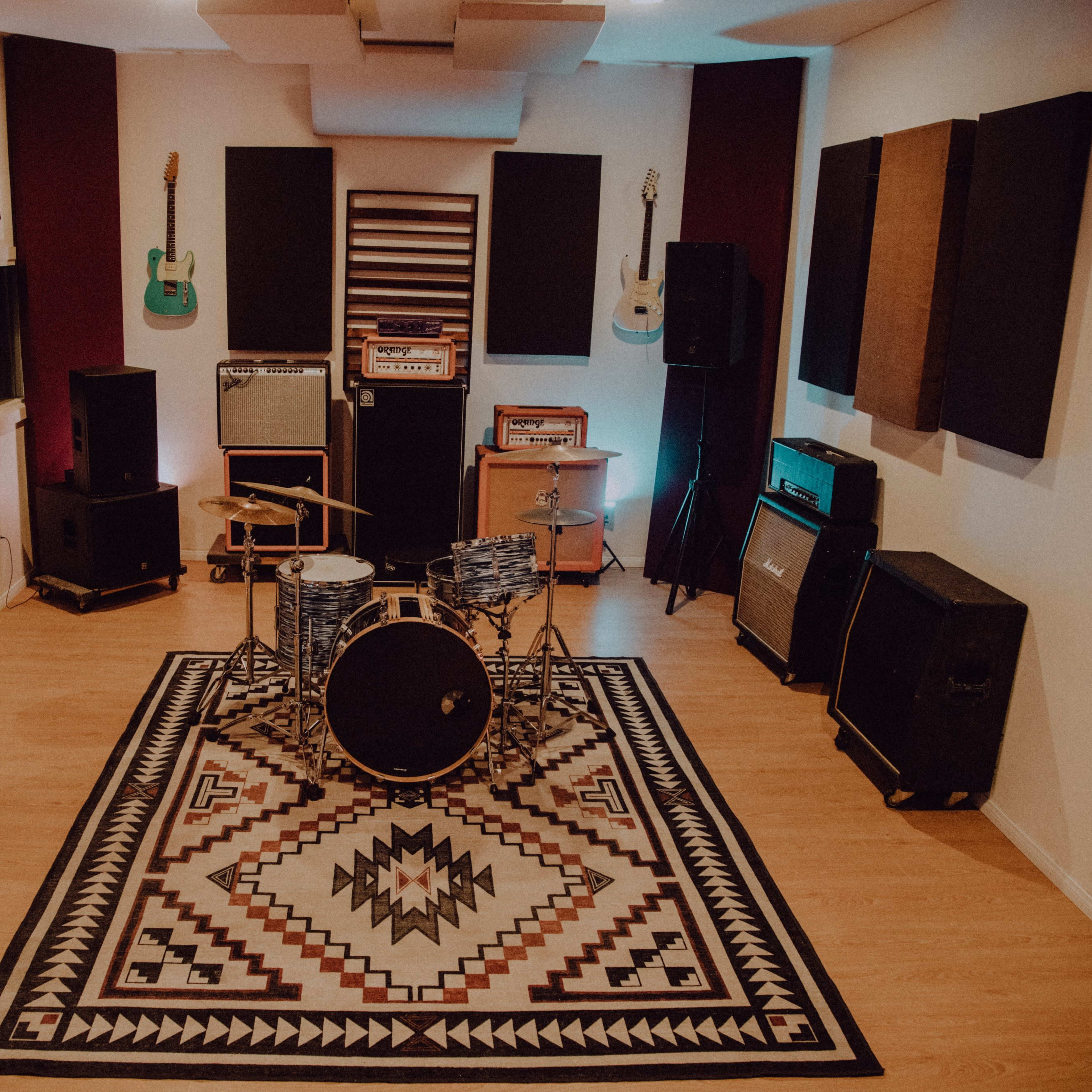 A music studio featuring a drum set in the center, surrounded by amplifiers and guitars mounted on the walls.