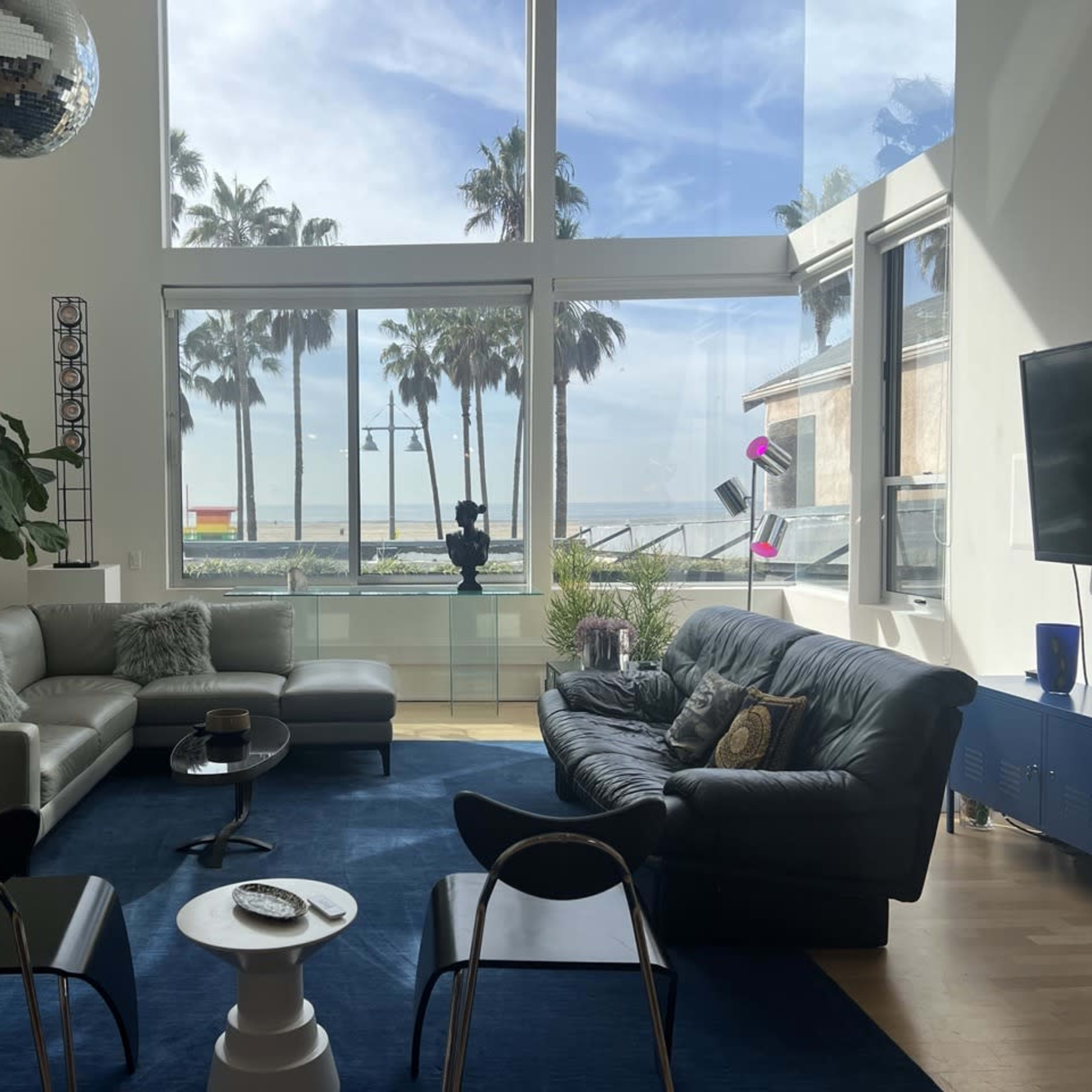 A modern living room features a large window showcasing palm trees and the ocean, with two sofas, a coffee table, and a disco ball hanging from the ceiling.