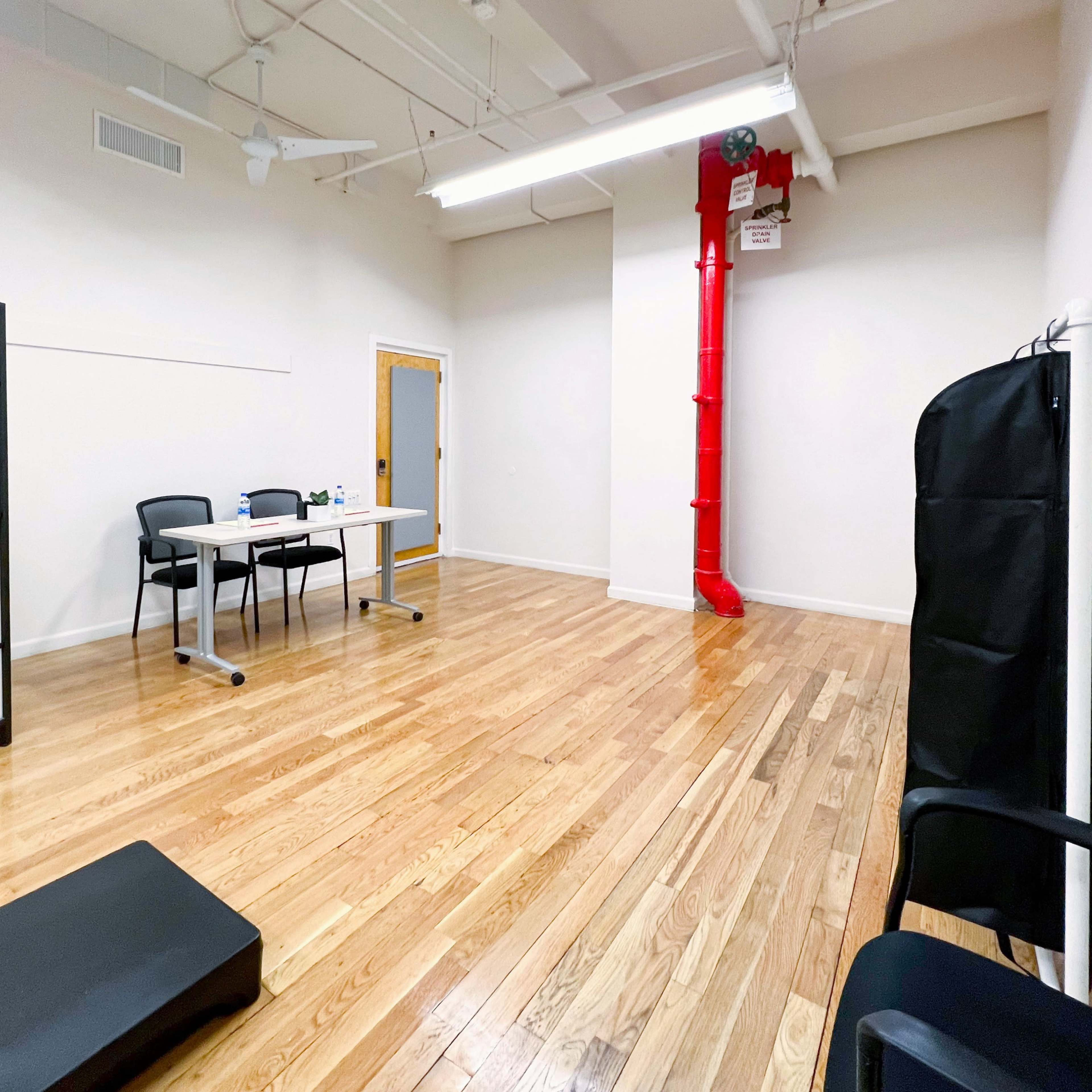 A sparse indoor room with wooden flooring, a folding screen, a table with chairs, and a red pipe on the wall.