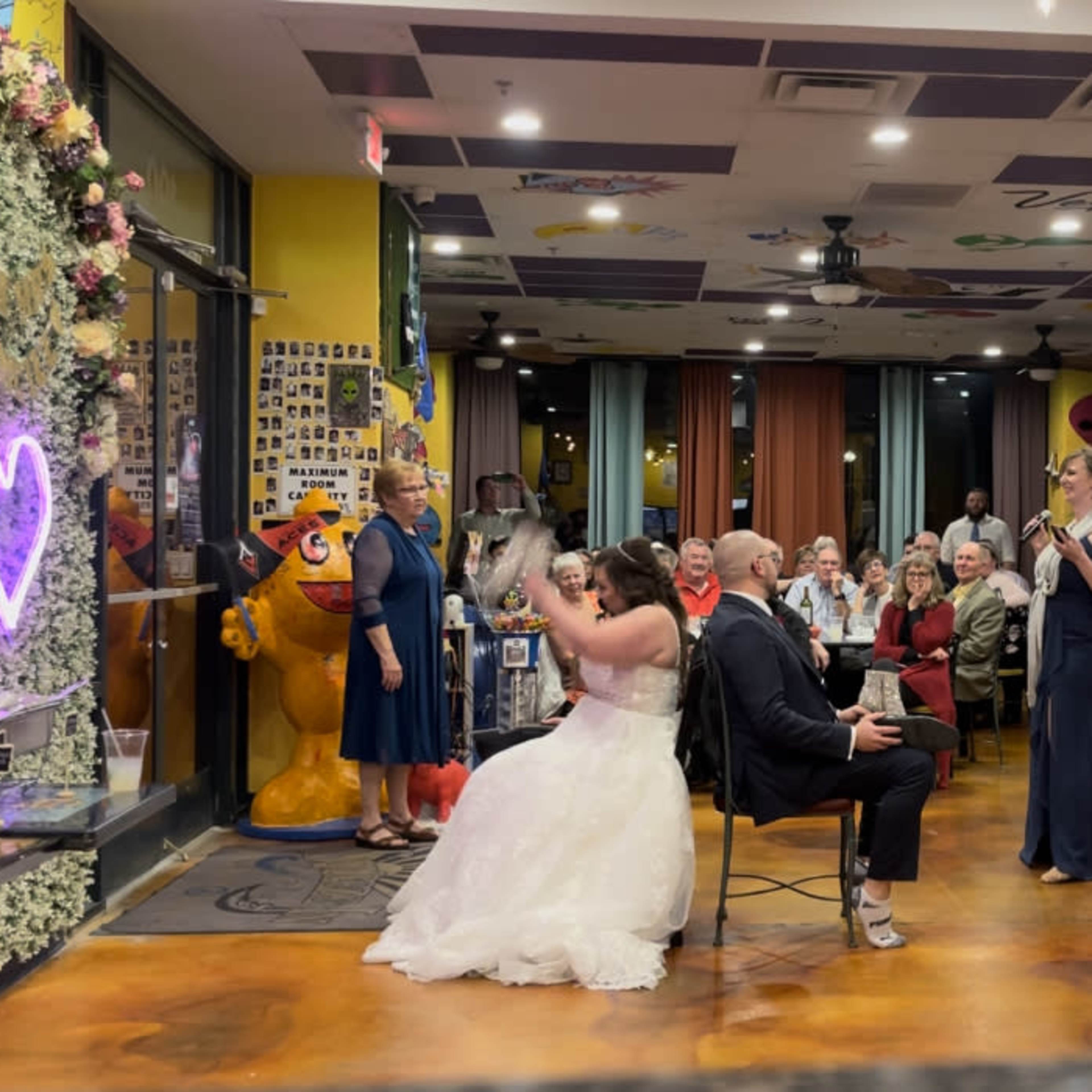 A bride in a white dress playfully tosses her bouquet while seated next to a groom at a decorated reception venue filled with guests.