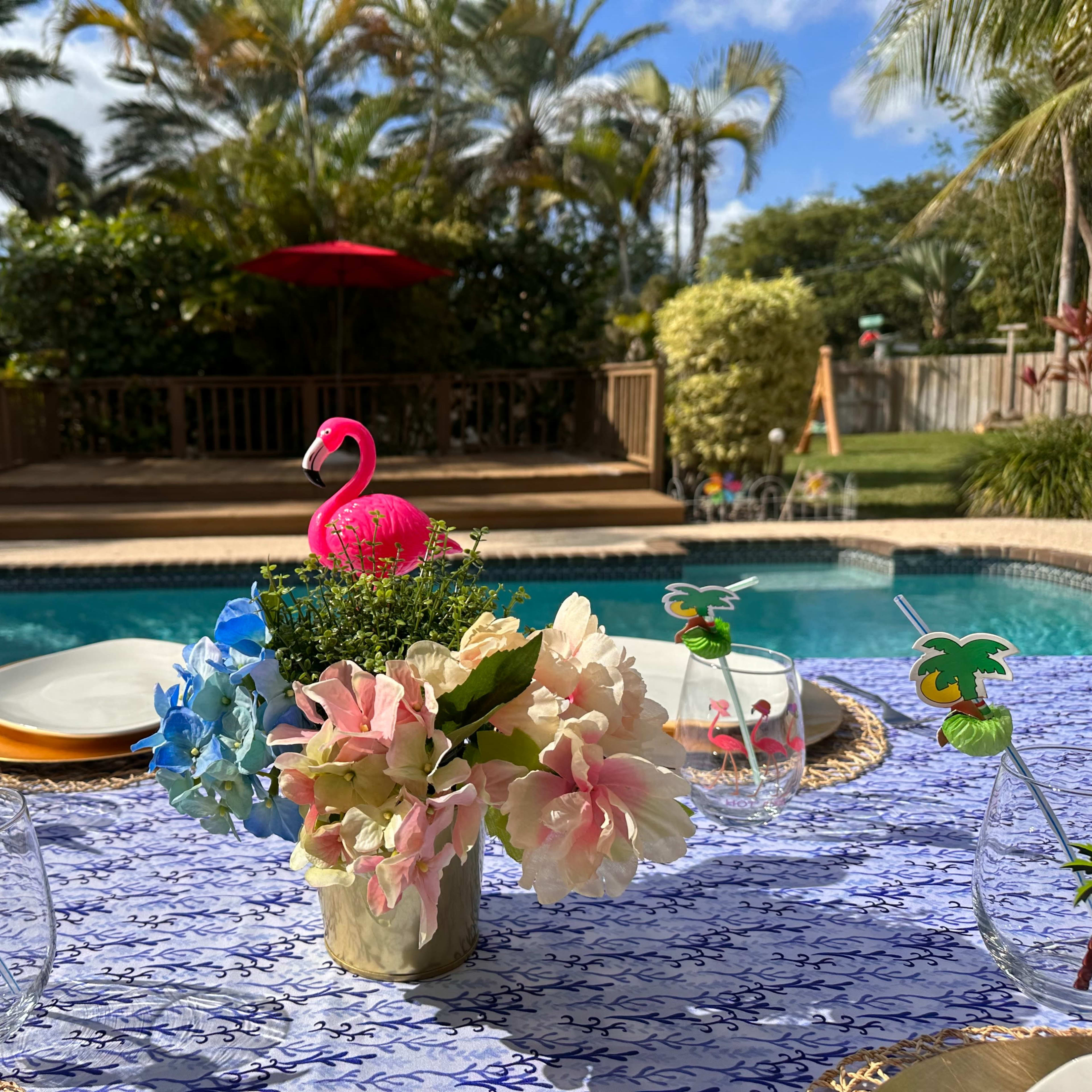 A table set with decorative glasses and a floral centerpiece overlooks a swimming pool and a deck surrounded by palm trees.