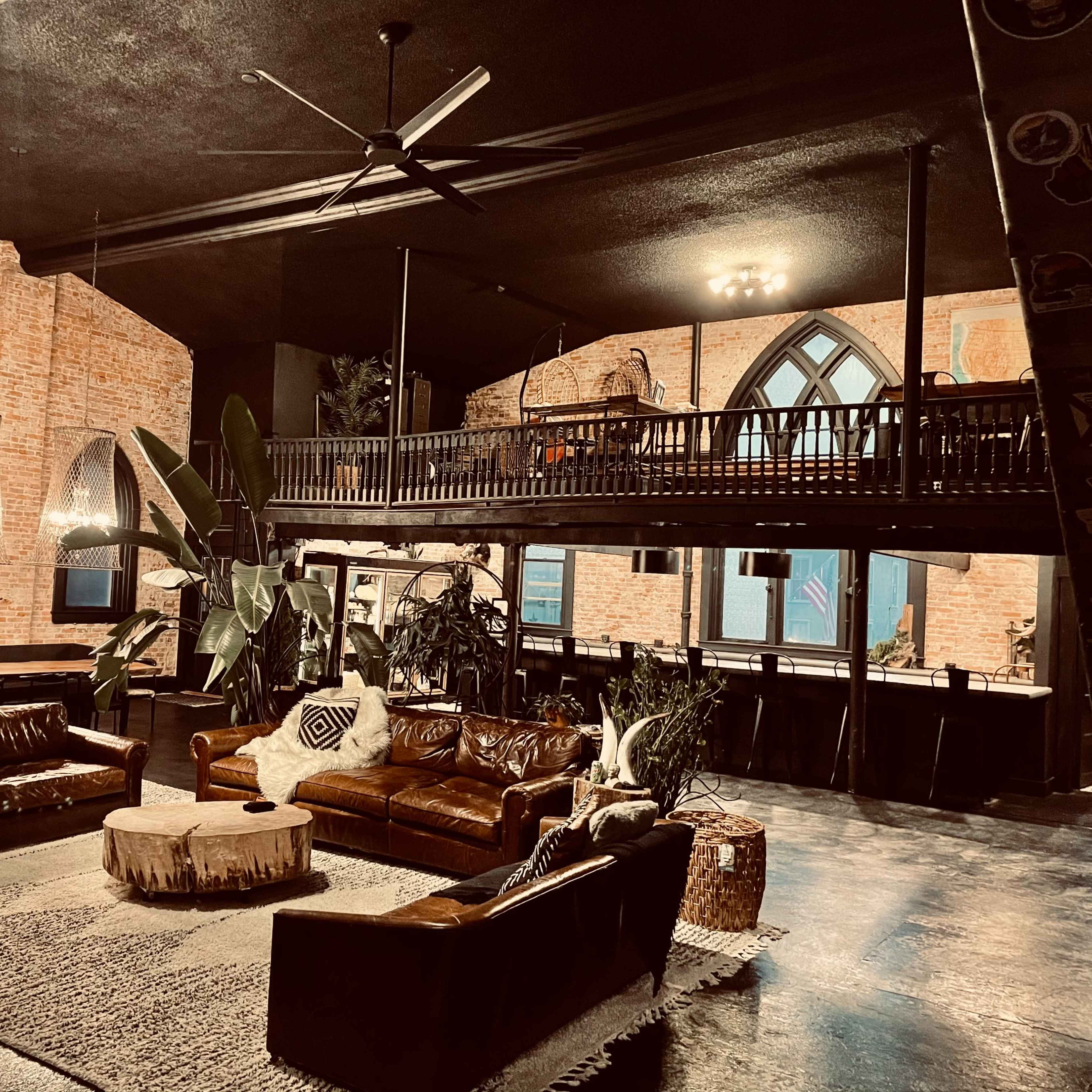 A spacious, industrial-style interior with two leather sofas, a round coffee table, large plants, and an upper loft area featuring gothic-style windows.