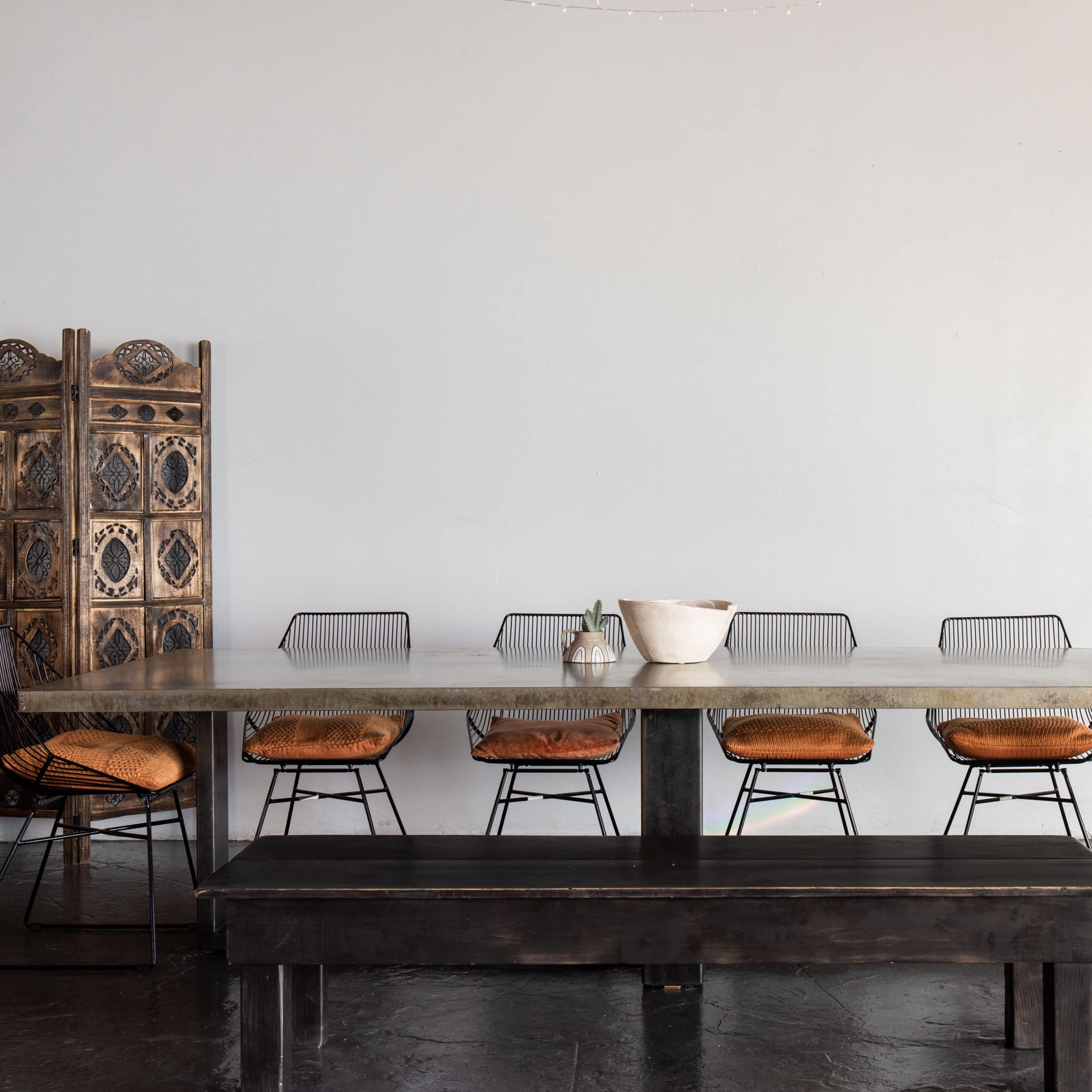 A large, rectangular dining table with metal chairs and a long bench is set against a plain wall, beside a decorative screen and a window.