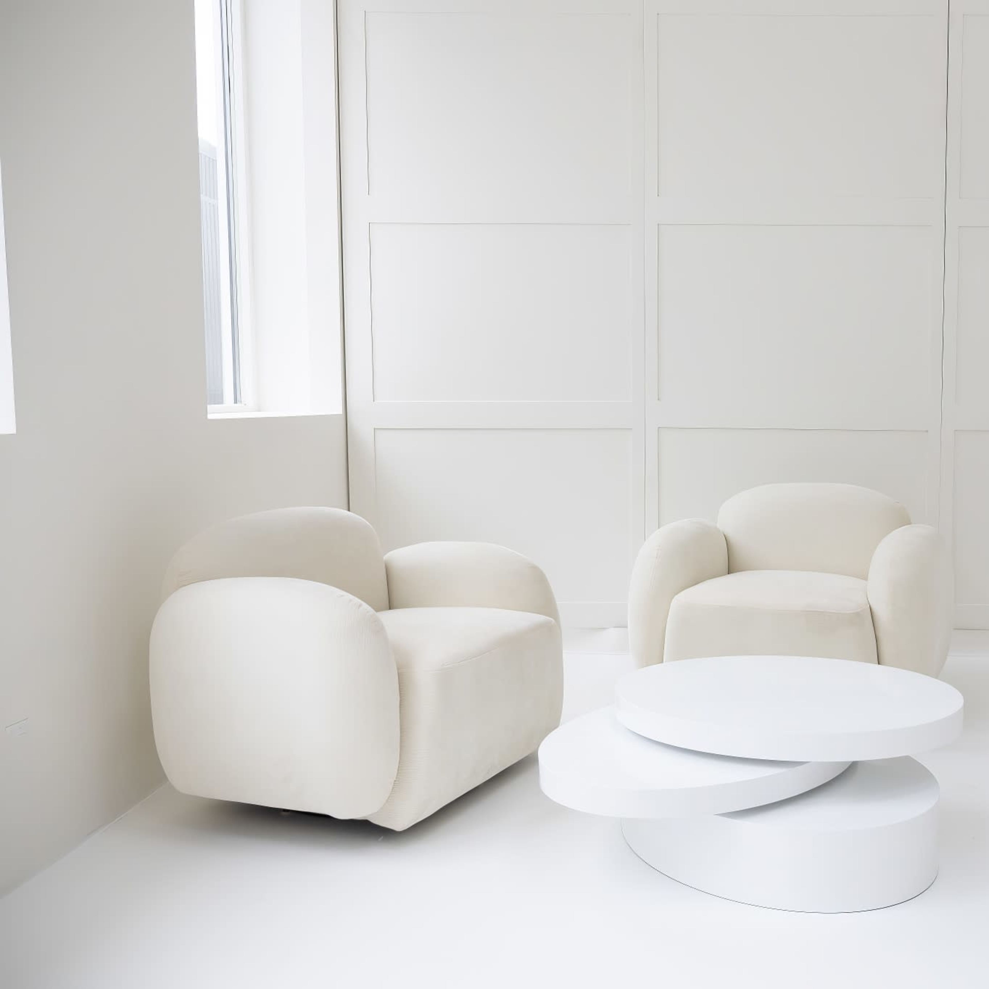 A minimalist interior with two cream-colored armchairs and a white modern coffee table against a plain wall.