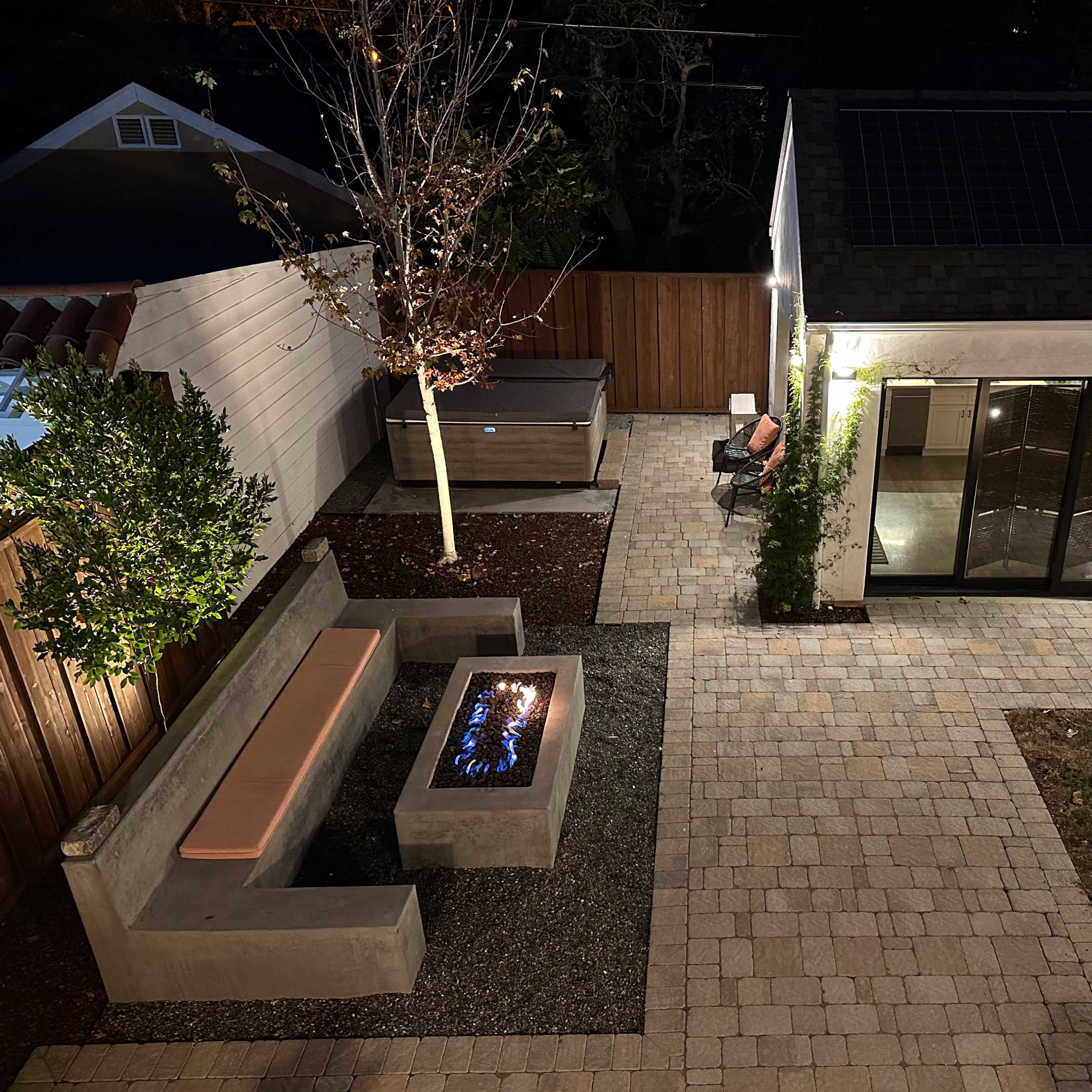 A modern outdoor patio featuring a stone fire pit, a concrete bench, and a seating area with patio chairs, surrounded by landscaping and illuminated at night.