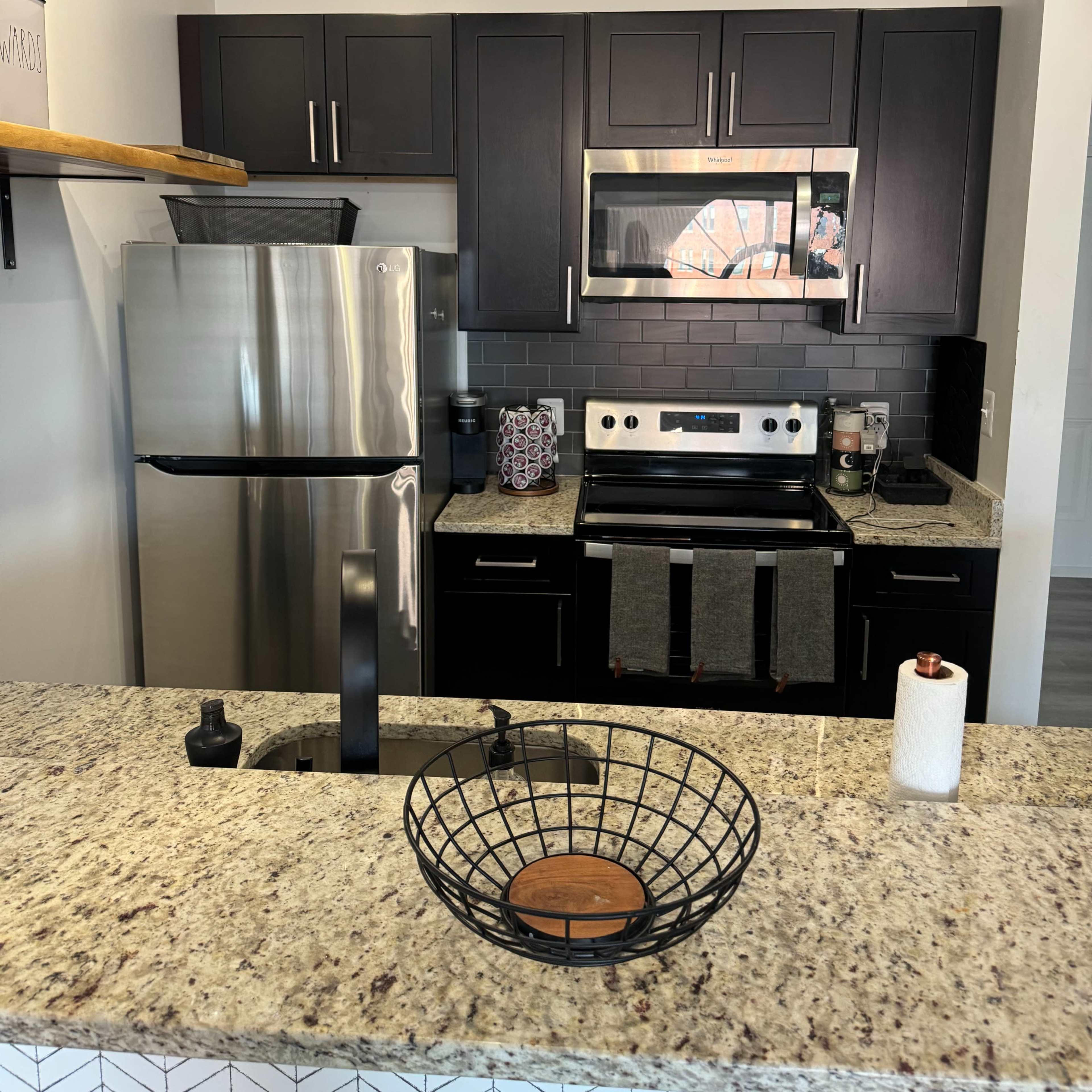 A modern kitchen with dark cabinetry, stainless steel appliances, and a granite countertop featuring a wire fruit bowl.