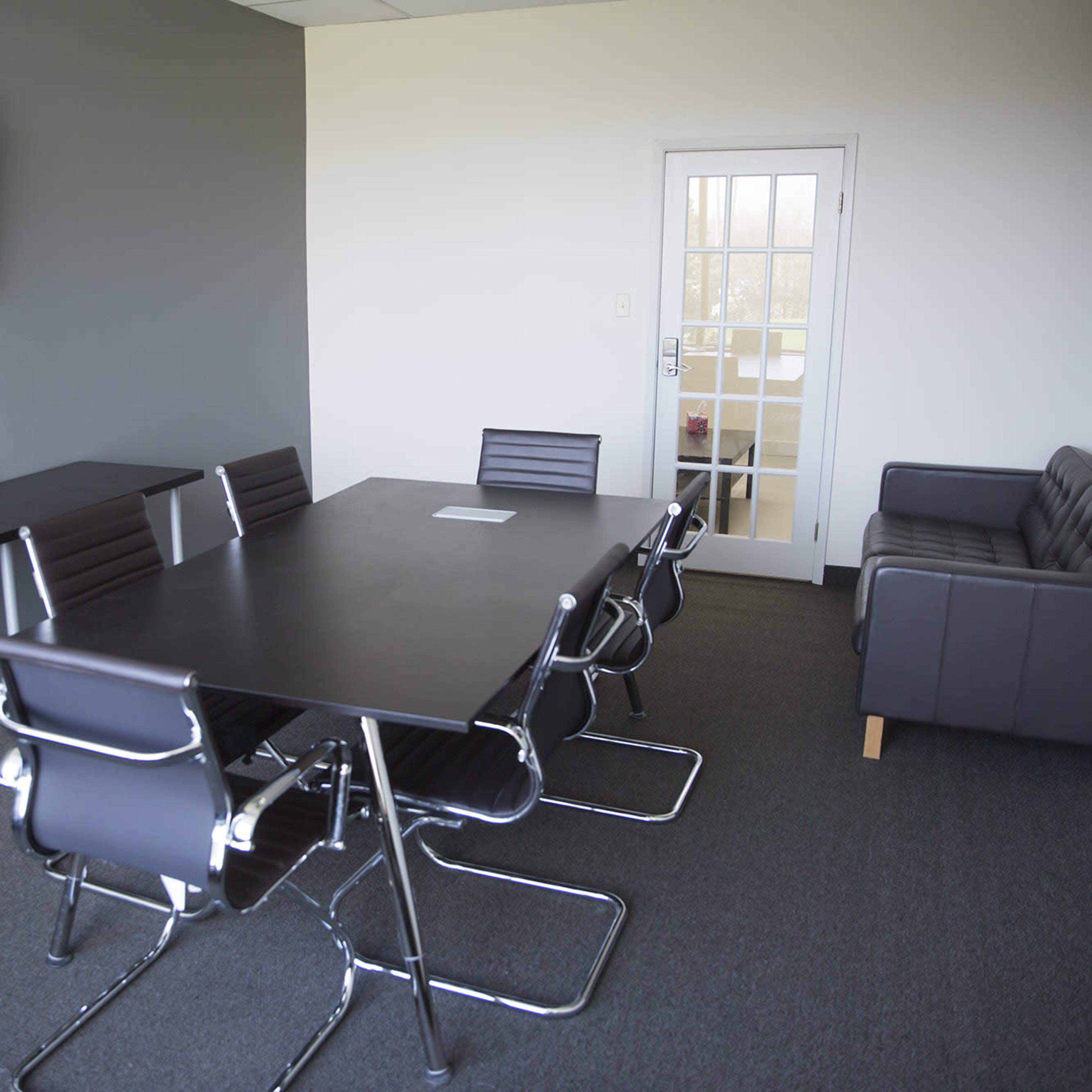 A modern conference room with a long table, several chairs, a television mounted on the wall, and a door leading to another area.