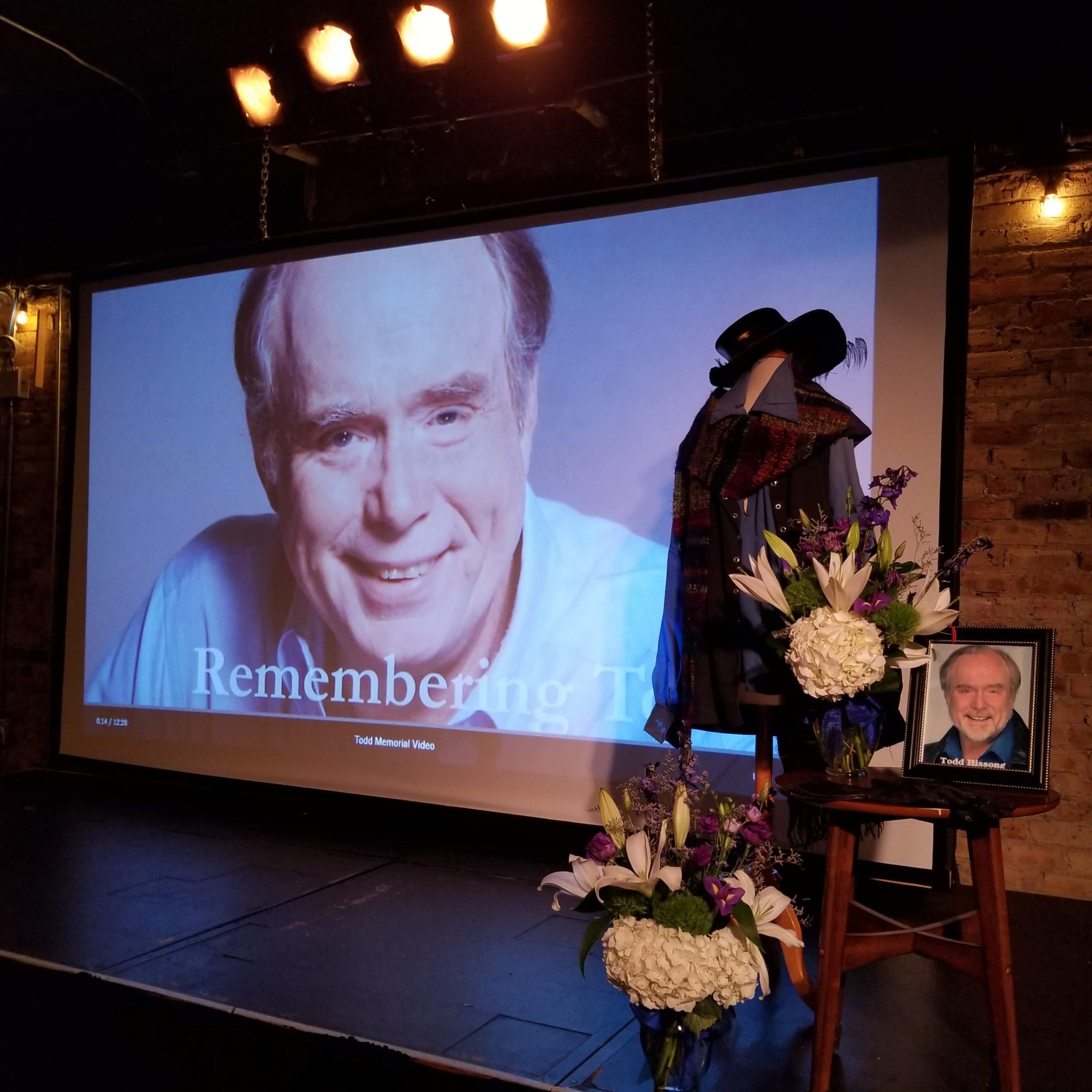 The scene features a stage setup with a large screen displaying "Remembering Them," alongside a framed photograph of a man, flowers, and a hat resting on a microphone stand.