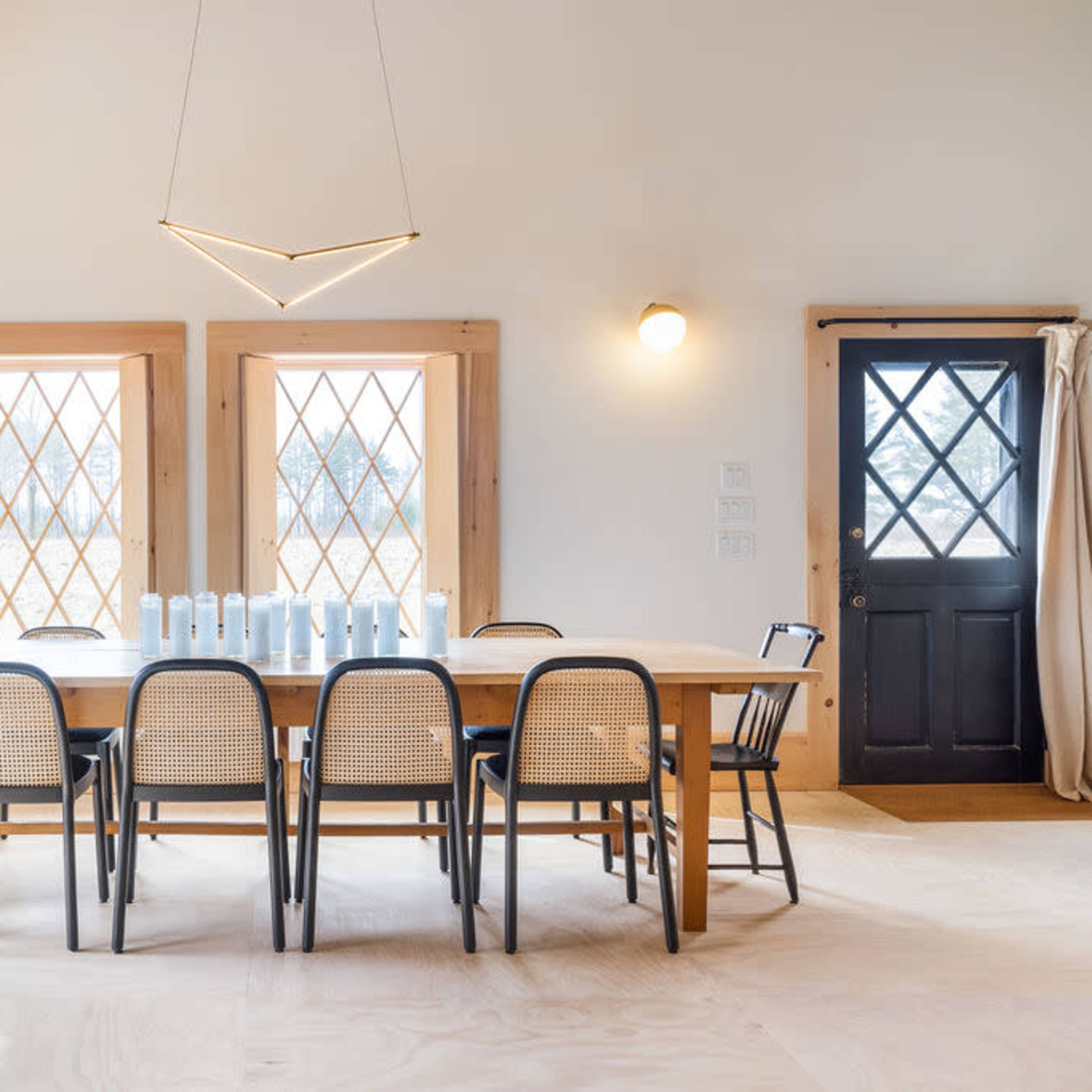 A modern dining room features a long wooden table surrounded by black and cane chairs, with large diamond-patterned windows and a black door.