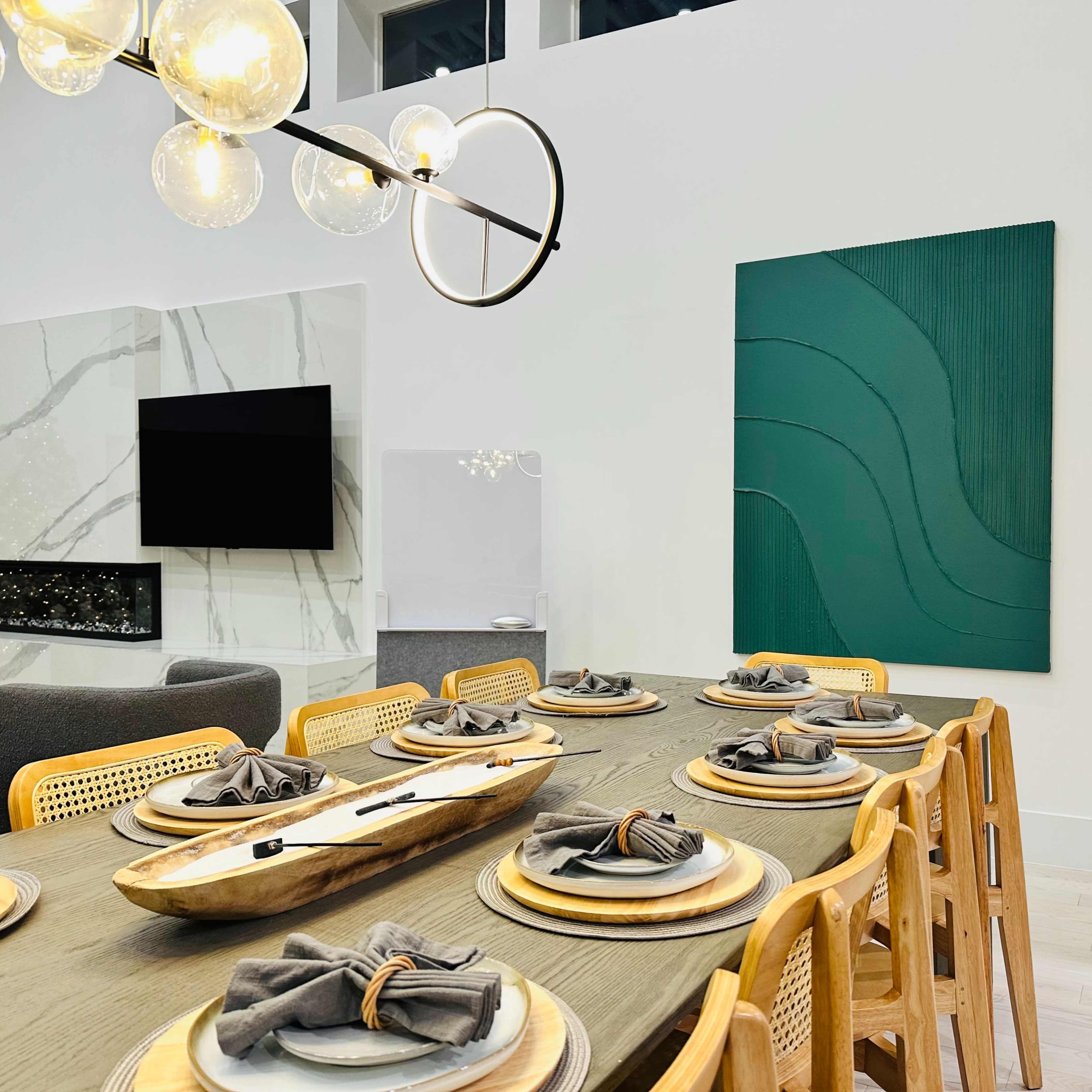 A modern dining room features a long wooden table set for eight, with a large abstract green artwork on the wall and a pendant light above.