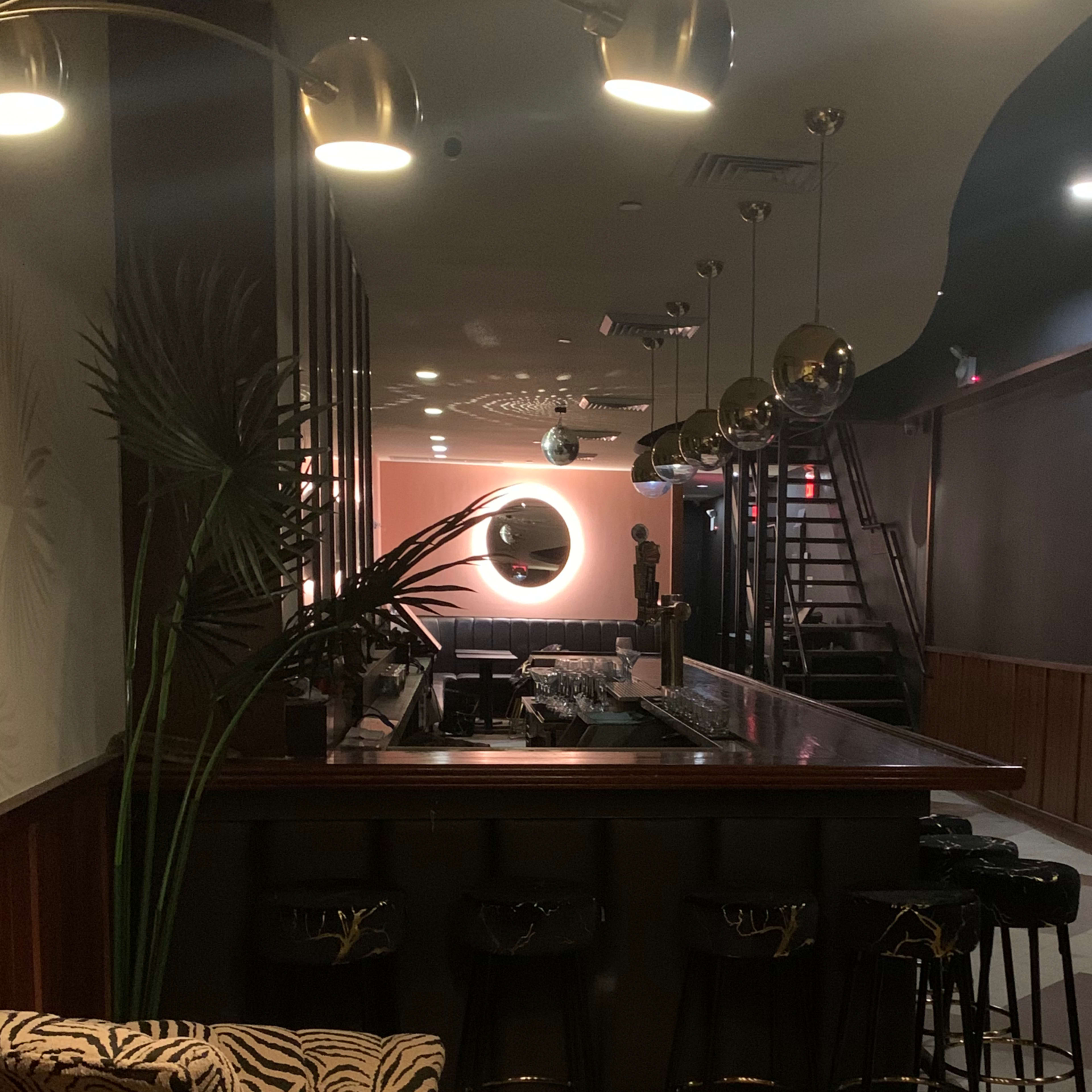 A dimly lit bar area with a counter, several bar stools, and stairs leading to an upper level.