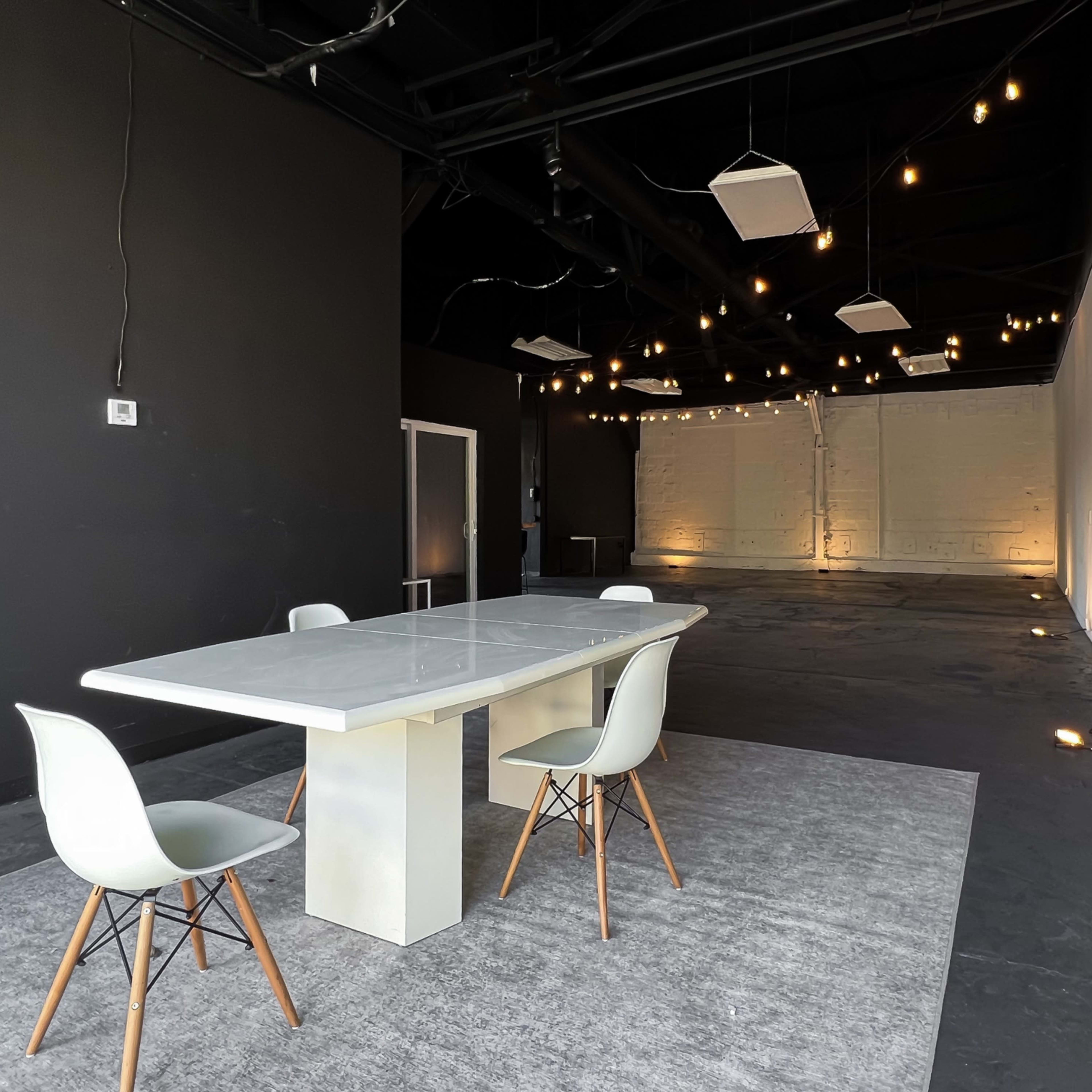 A large, minimalist meeting space features a rectangular table surrounded by four chairs, with soft lighting highlighting the walls.