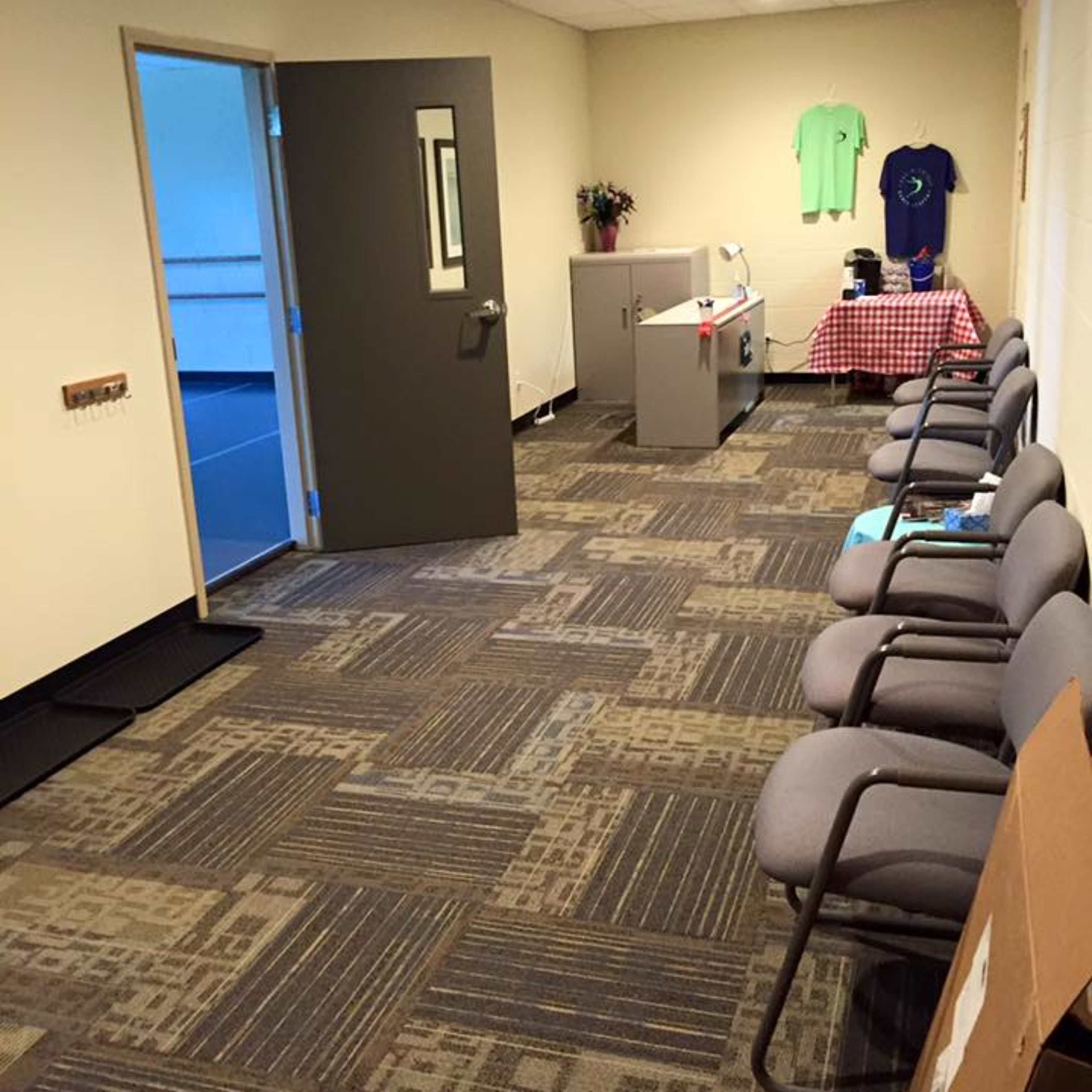 A hallway with gray chairs along one side, a desk with a checkered tablecloth at the end, and a door leading to another room.