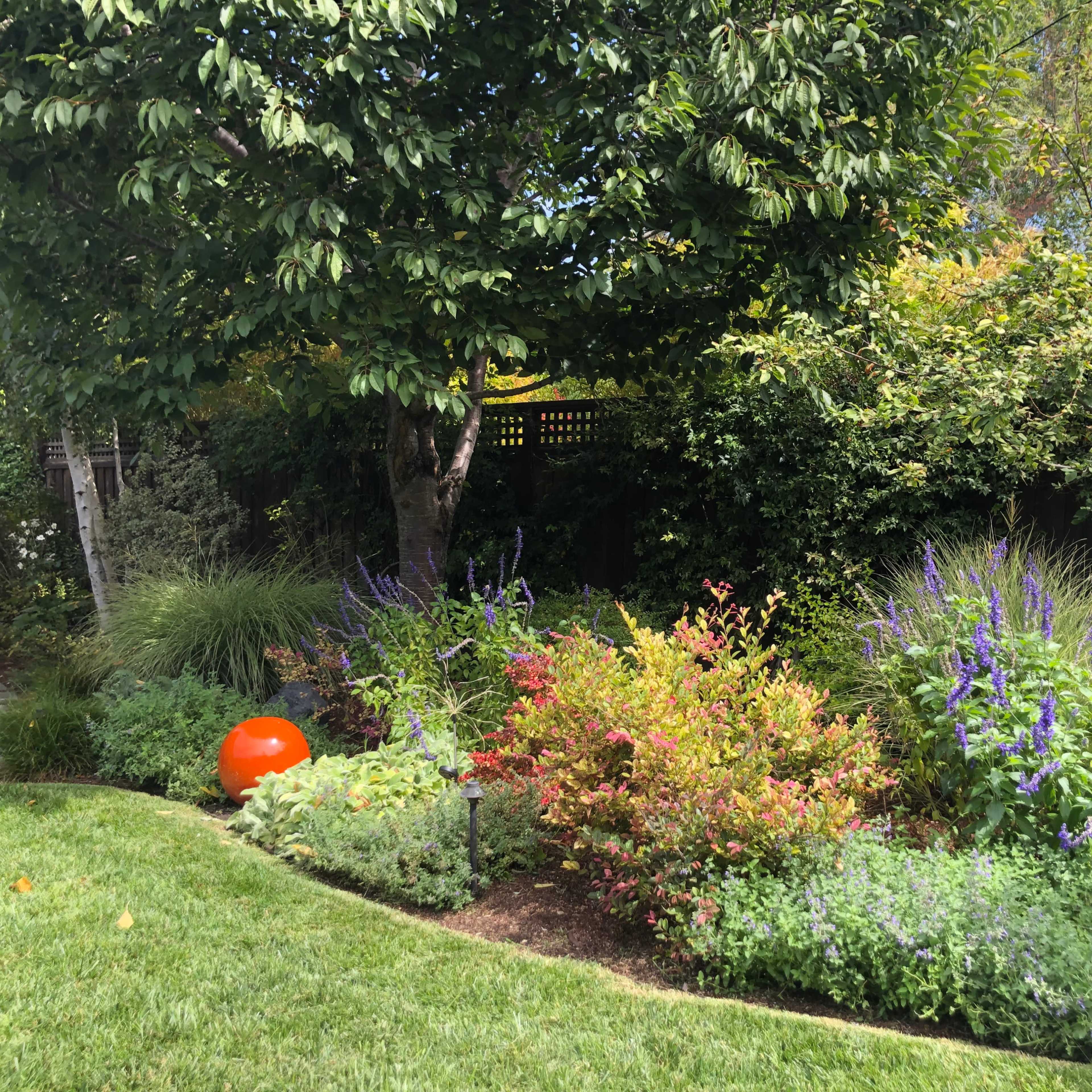 A circular orange sculpture sits among vibrant flowerbeds and lush greenery in a garden.