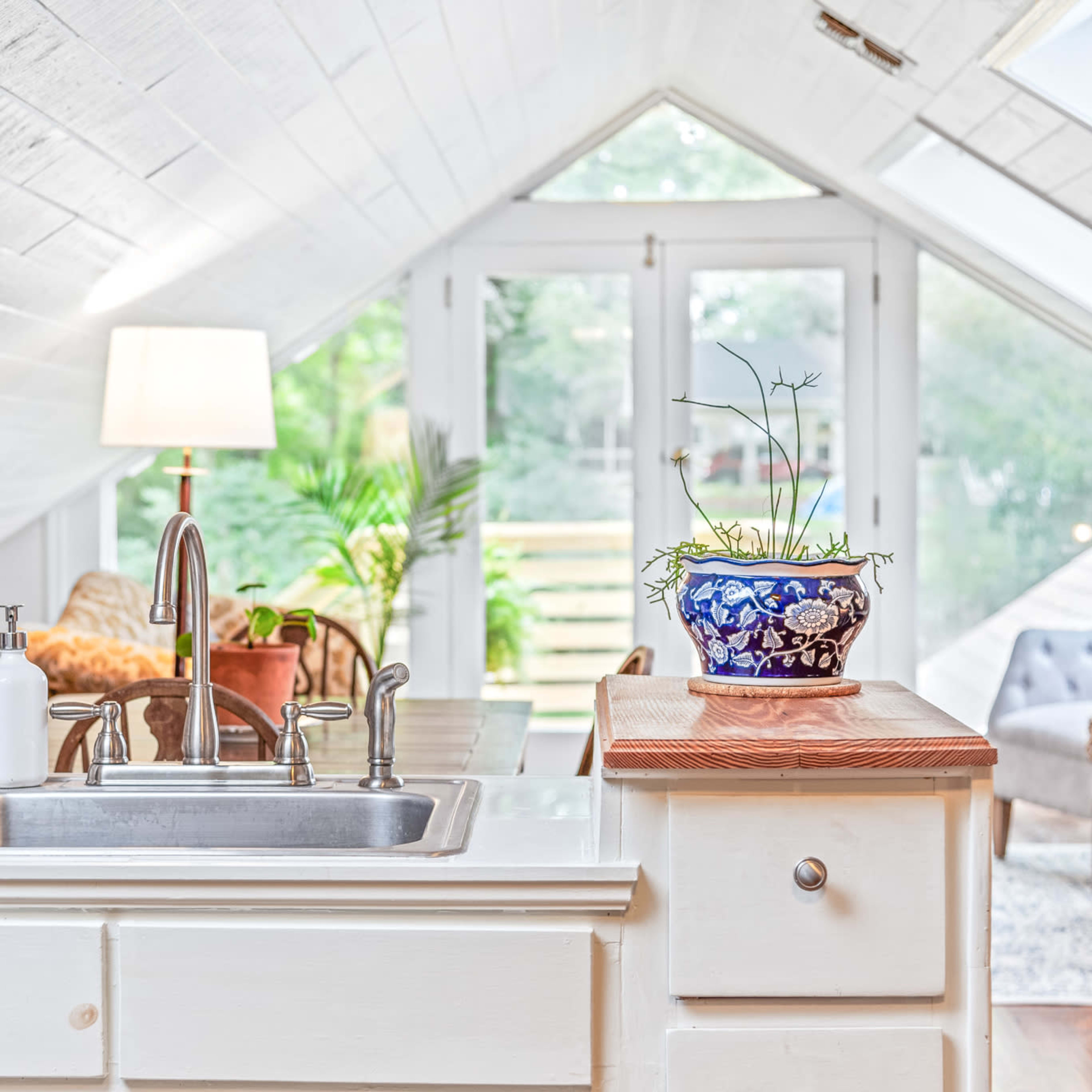 A bright, airy attic kitchen features a stainless steel sink, a decorative plant on the countertop, and large windows that bring in natural light.