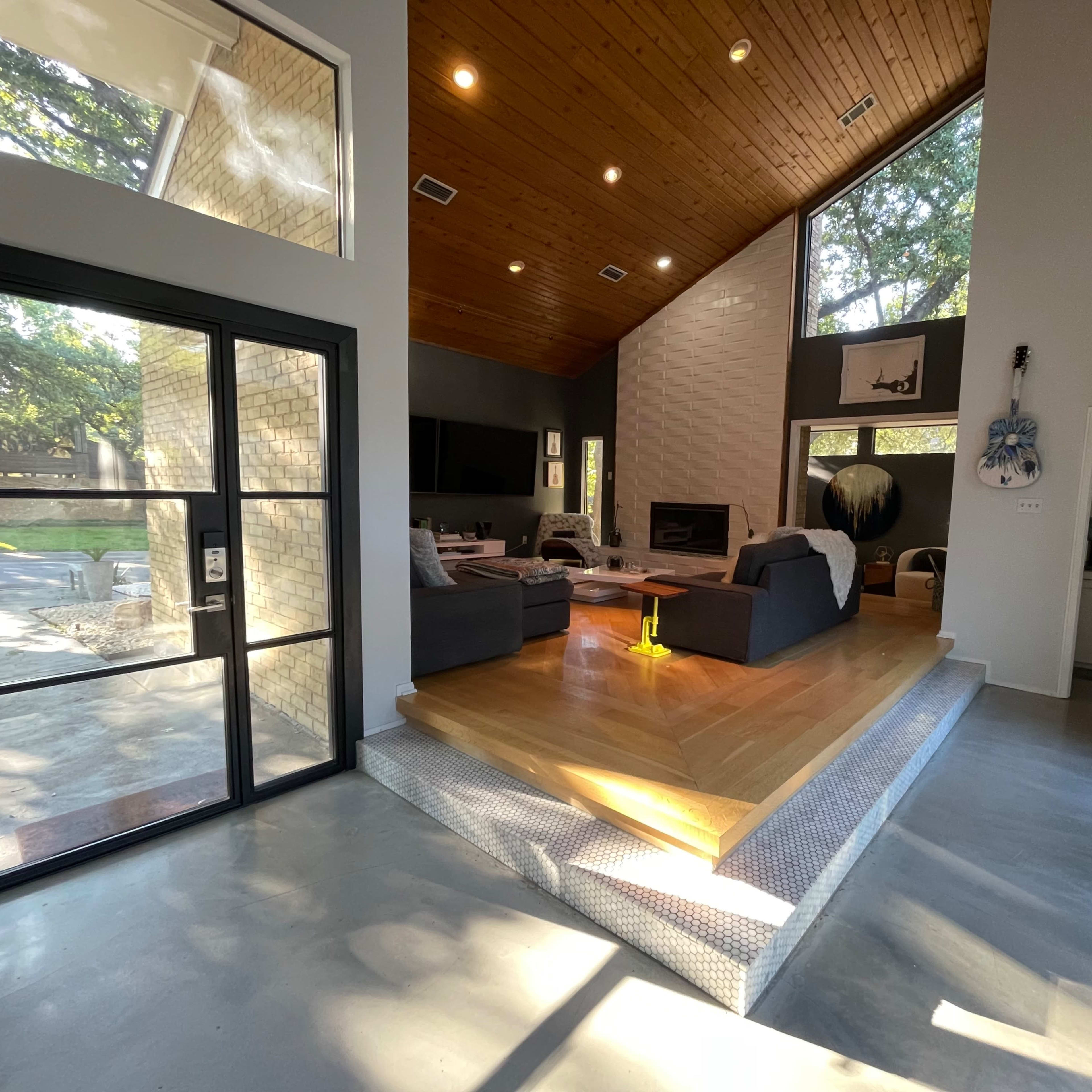 A modern living room with a vaulted wooden ceiling, large windows, and a sunken seating area featuring a fireplace.