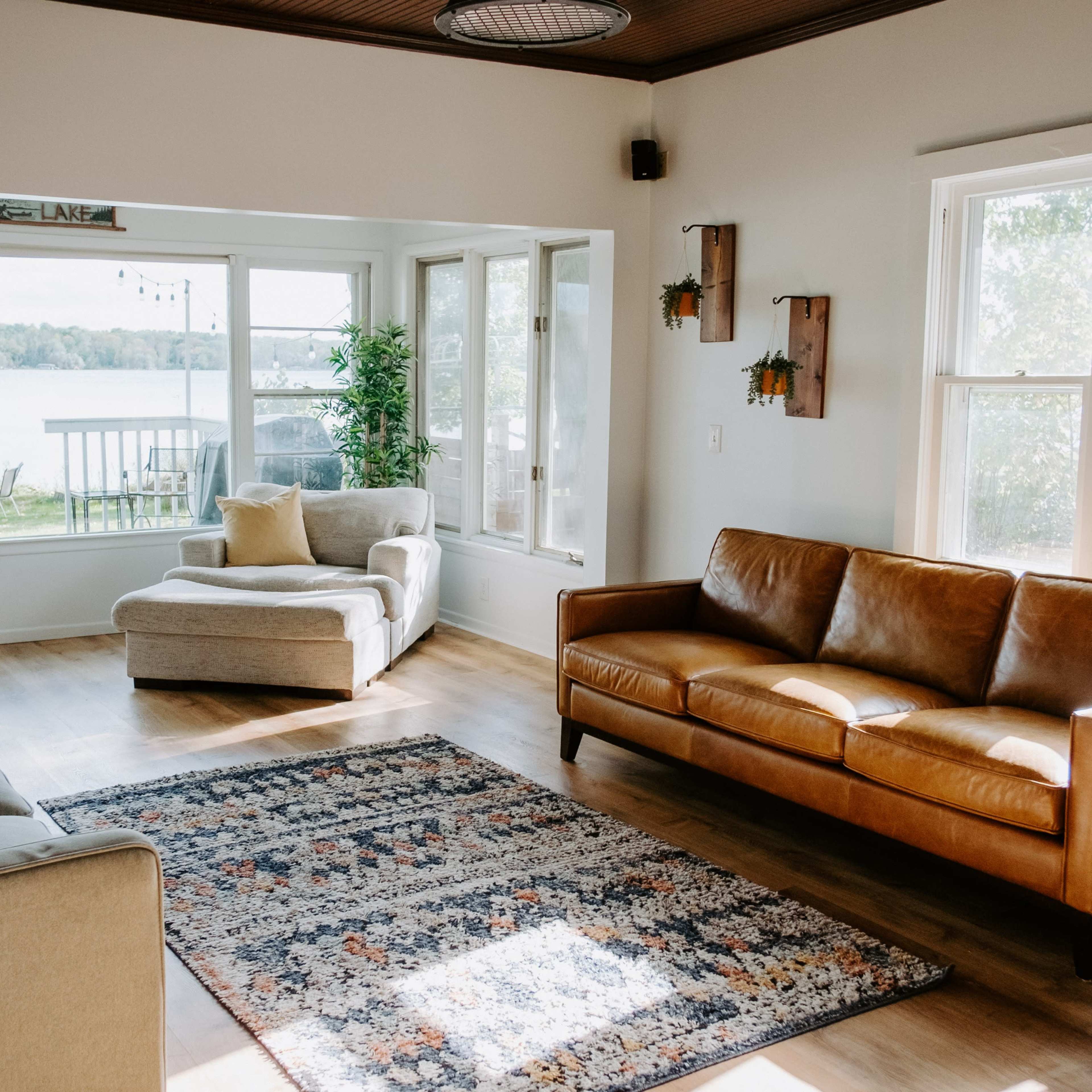 The living room features a beige sofa and a brown leather couch, with a patterned area rug on the wooden floor and large windows overlooking a lake.