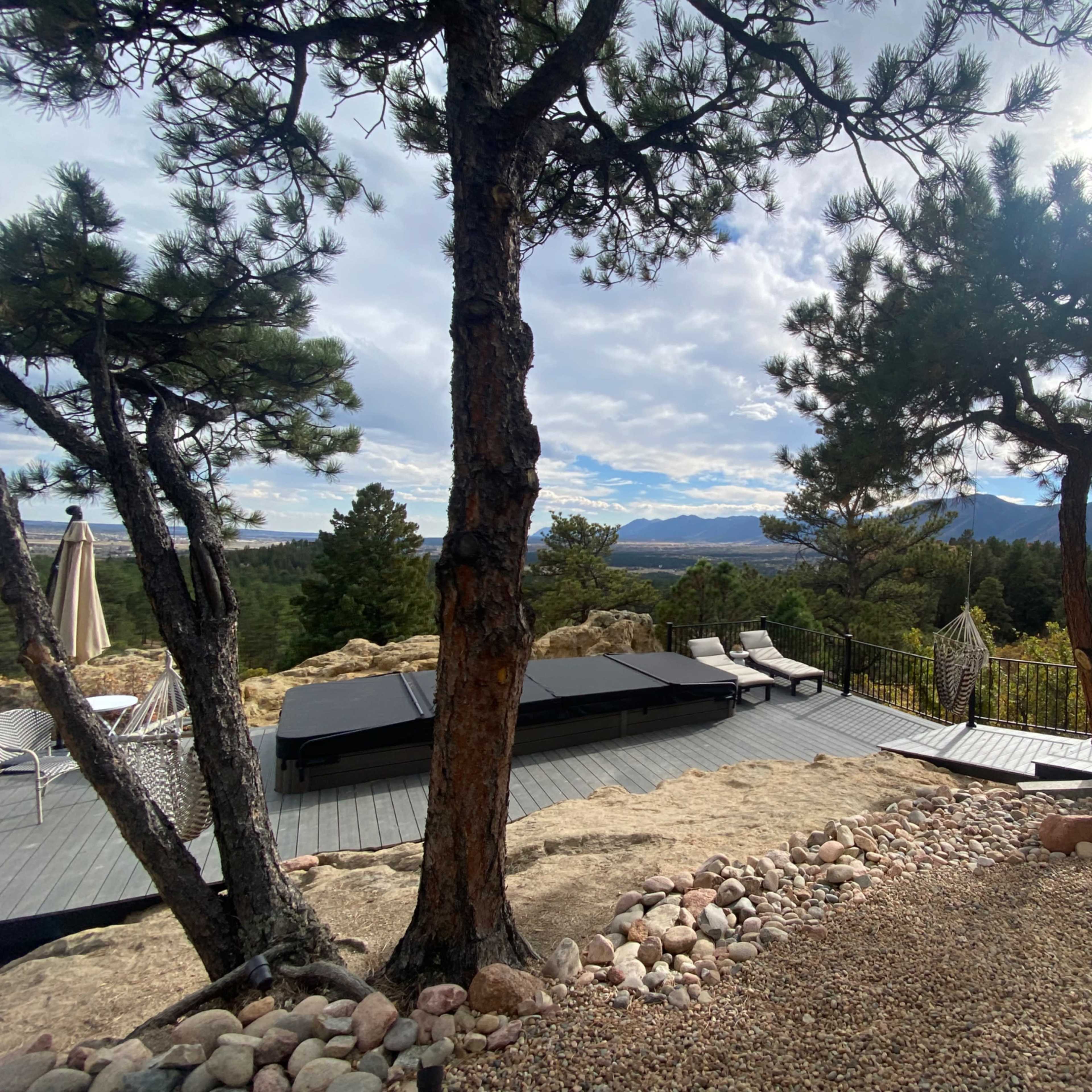 The scene features a patio area with lounge chairs, rocky landscaping, and a view of mountains and trees under a partly cloudy sky.