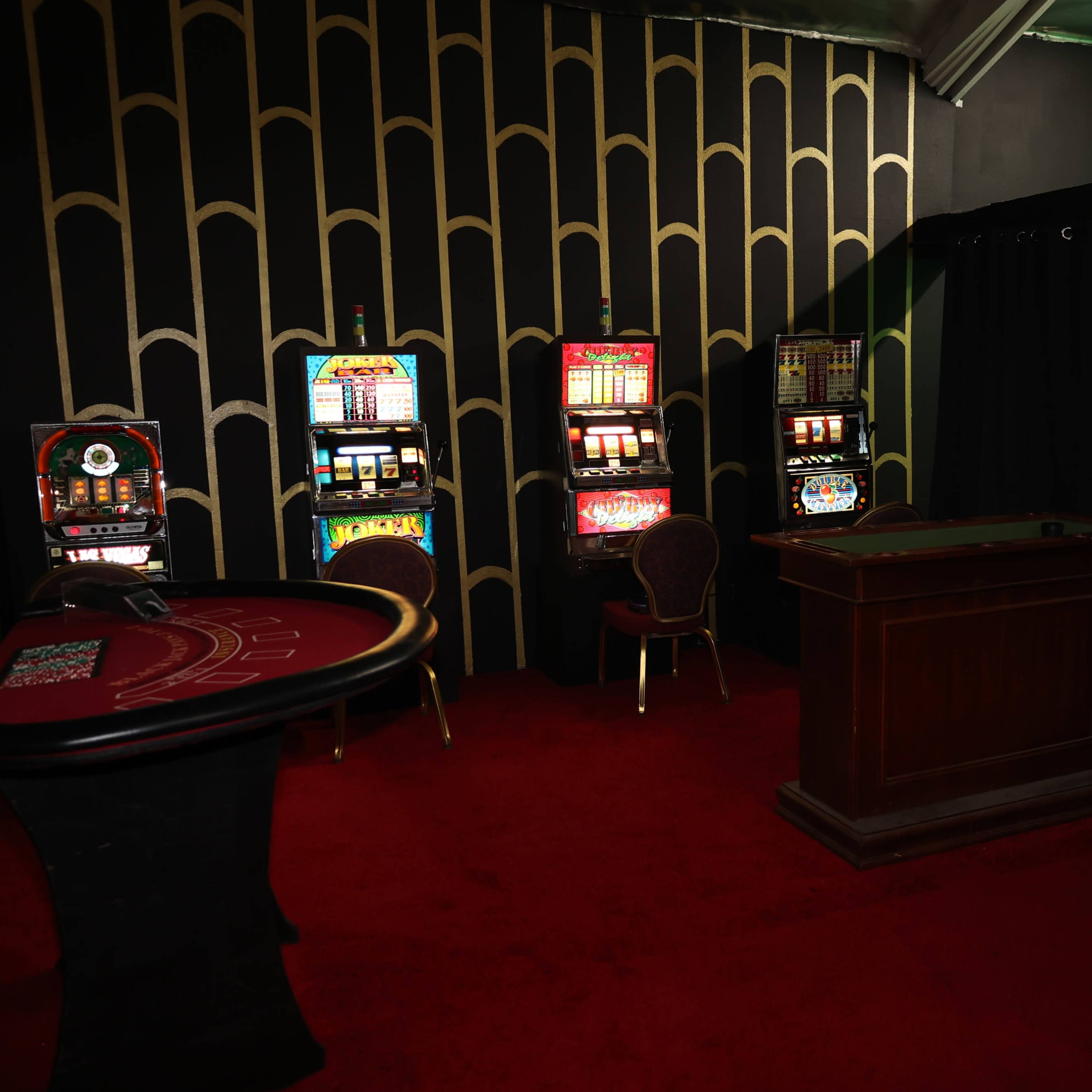A dimly lit gambling area with a red carpet, a poker table, and several slot machines against a patterned black wall.