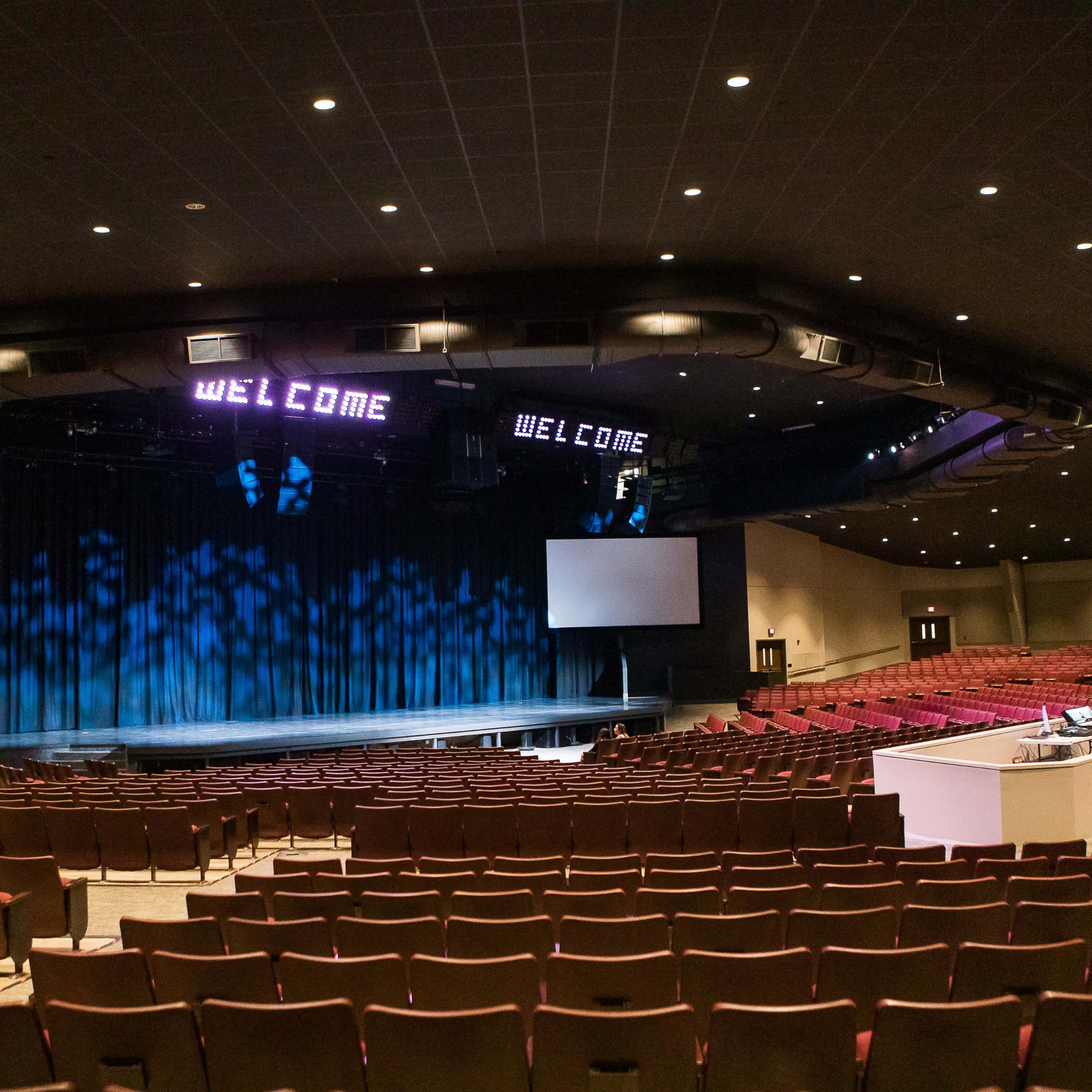 A spacious auditorium with a dark stage, a large blue curtain, and two signs that say "WELCOME" illuminated above the stage.