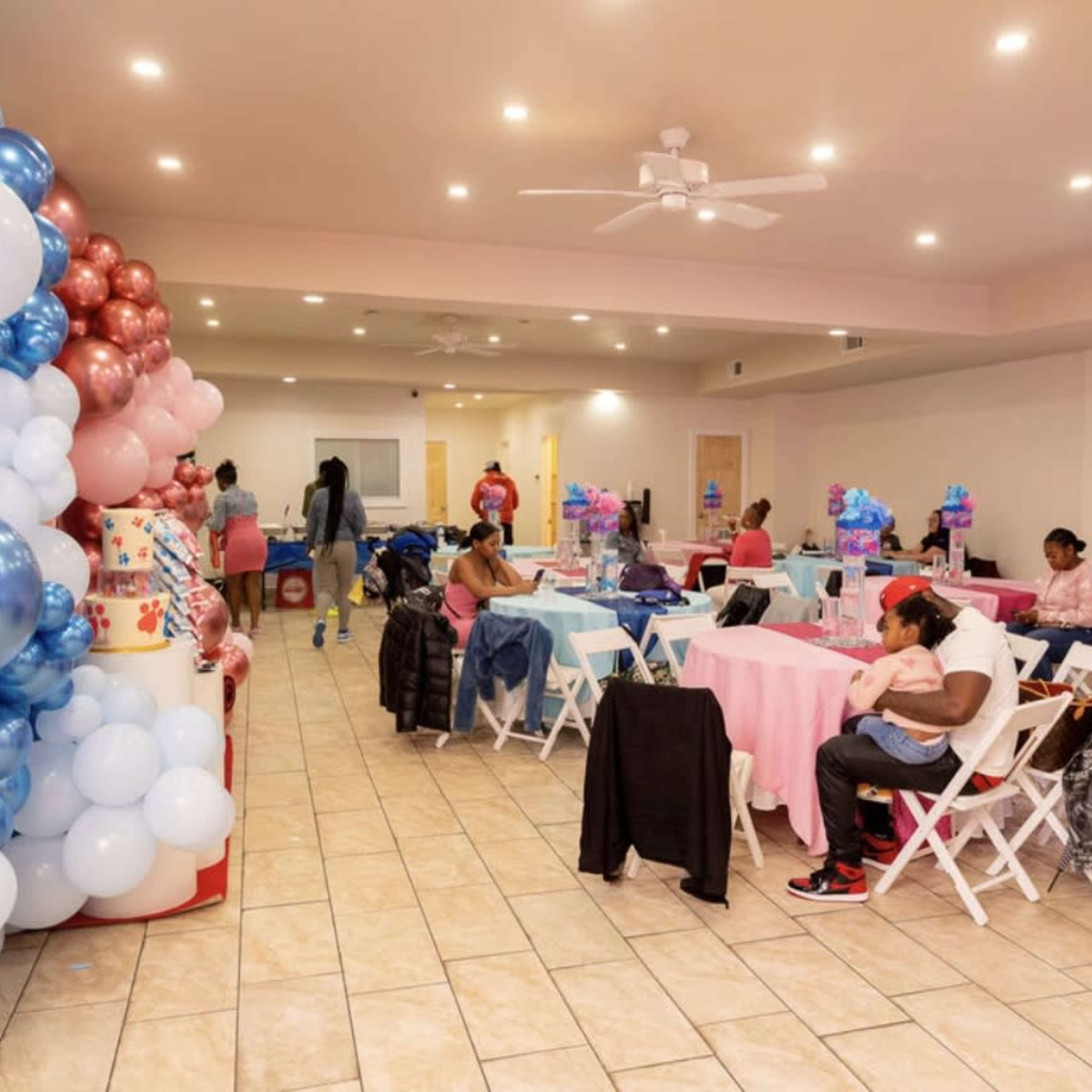 A decorated event space with tables set in pastel colors and a balloon arch in one corner, while guests engage in various activities.